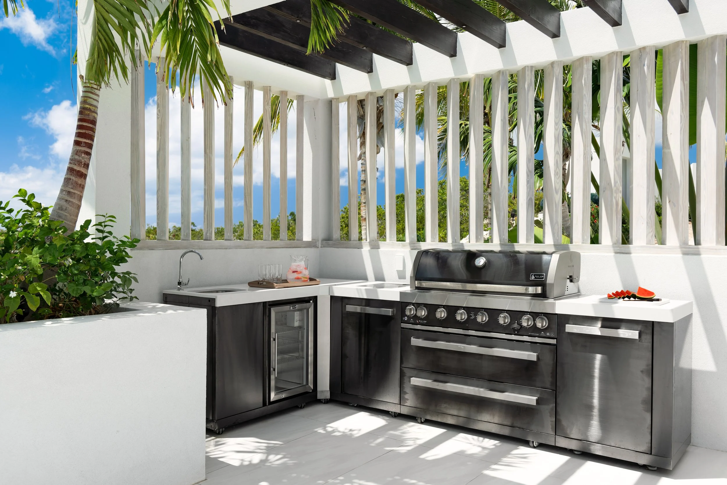 Outdoor kitchen with stainless steel grill, sink, and mini refrigerator, surrounded by white walls and fencing, with tropical trees and blue sky in the background.
