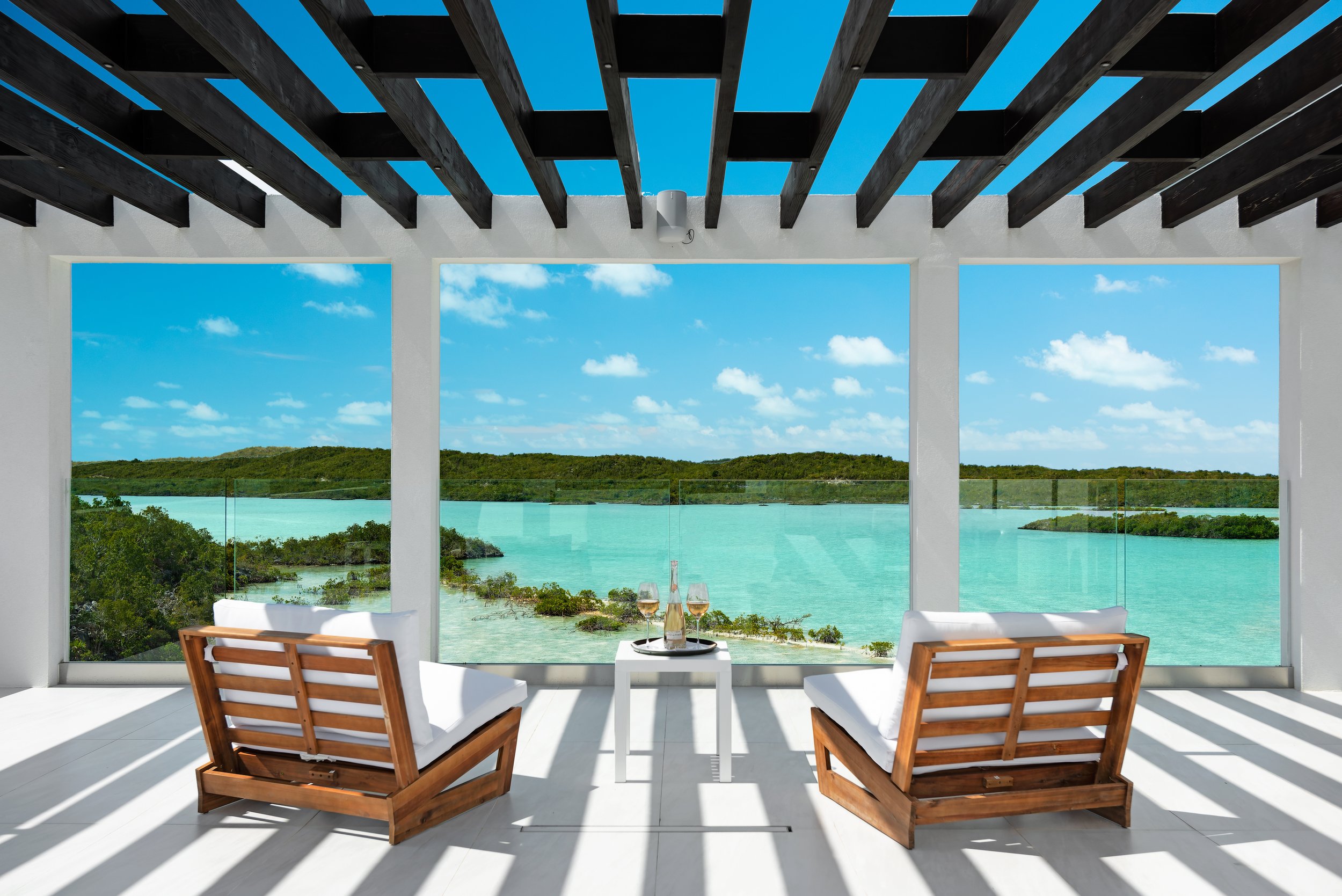 A modern indoor space with large glass windows overlooking a turquoise water body, green islands, and a partly cloudy sky. Two wooden chairs with white cushions face each other with a small white table between them, holding a bottle and two glasses o