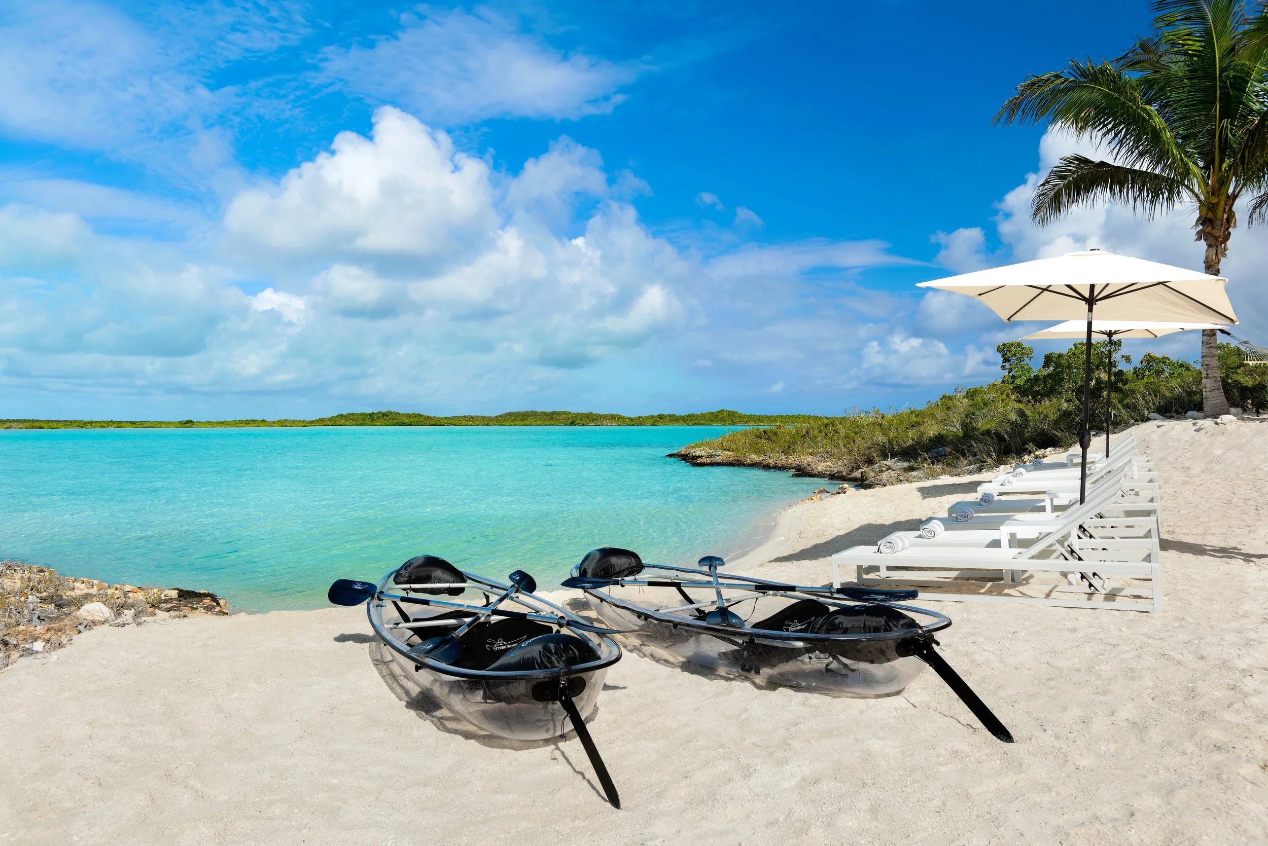Two kayaks on a sandy beach facing a turquoise water ocean with white lounge chairs, umbrellas, palm trees, and a blue sky with clouds.