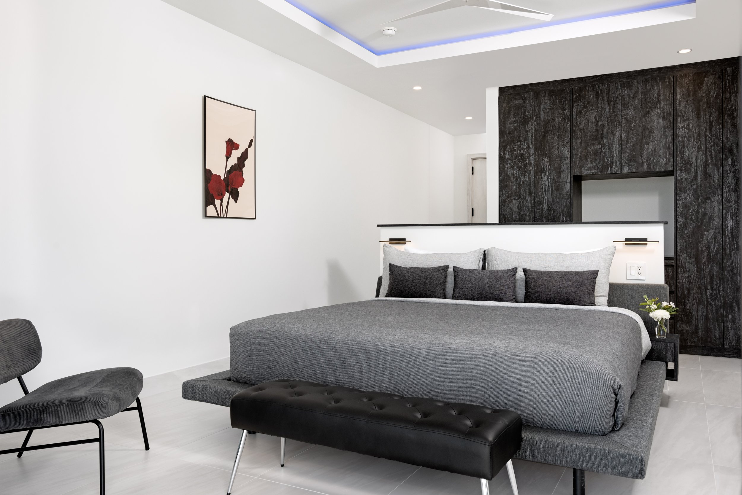 Modern bedroom with gray bed, black Leather Ottoman, gray chairs, white walls, dark wooden accent wall, and a wall art of red flowers.