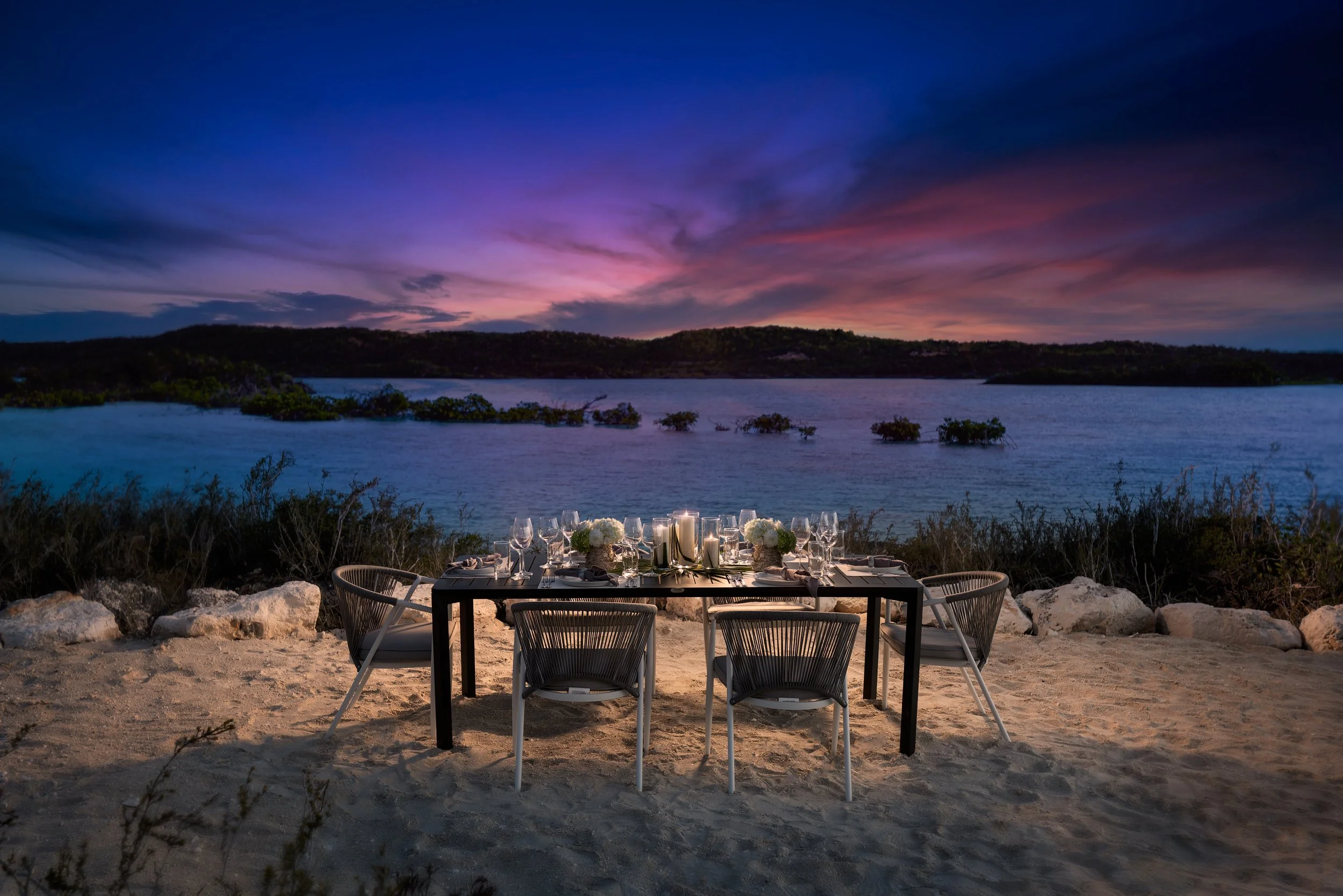 A dinner setup with a black rectangular table, white chairs, and elegant tableware on a sandy beach during sunset, with a lake and colorful sky in the background.