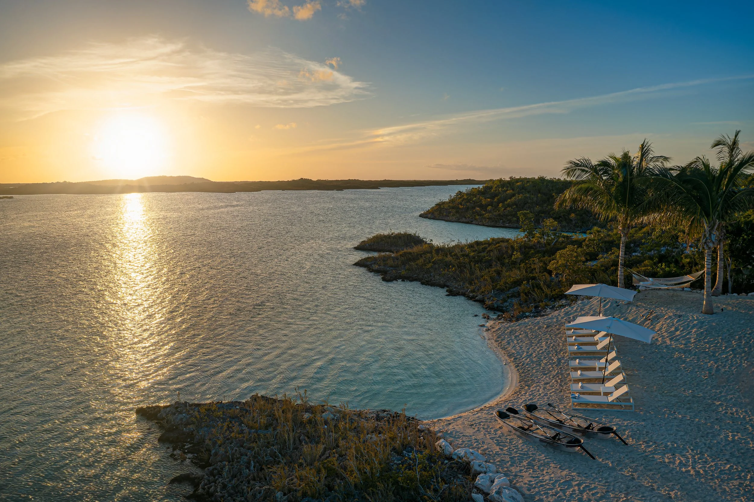 Sunset over a calm coastal bay with white sandy beach, beach chairs under umbrellas, paddle boats on the shore, and palm trees on the right.