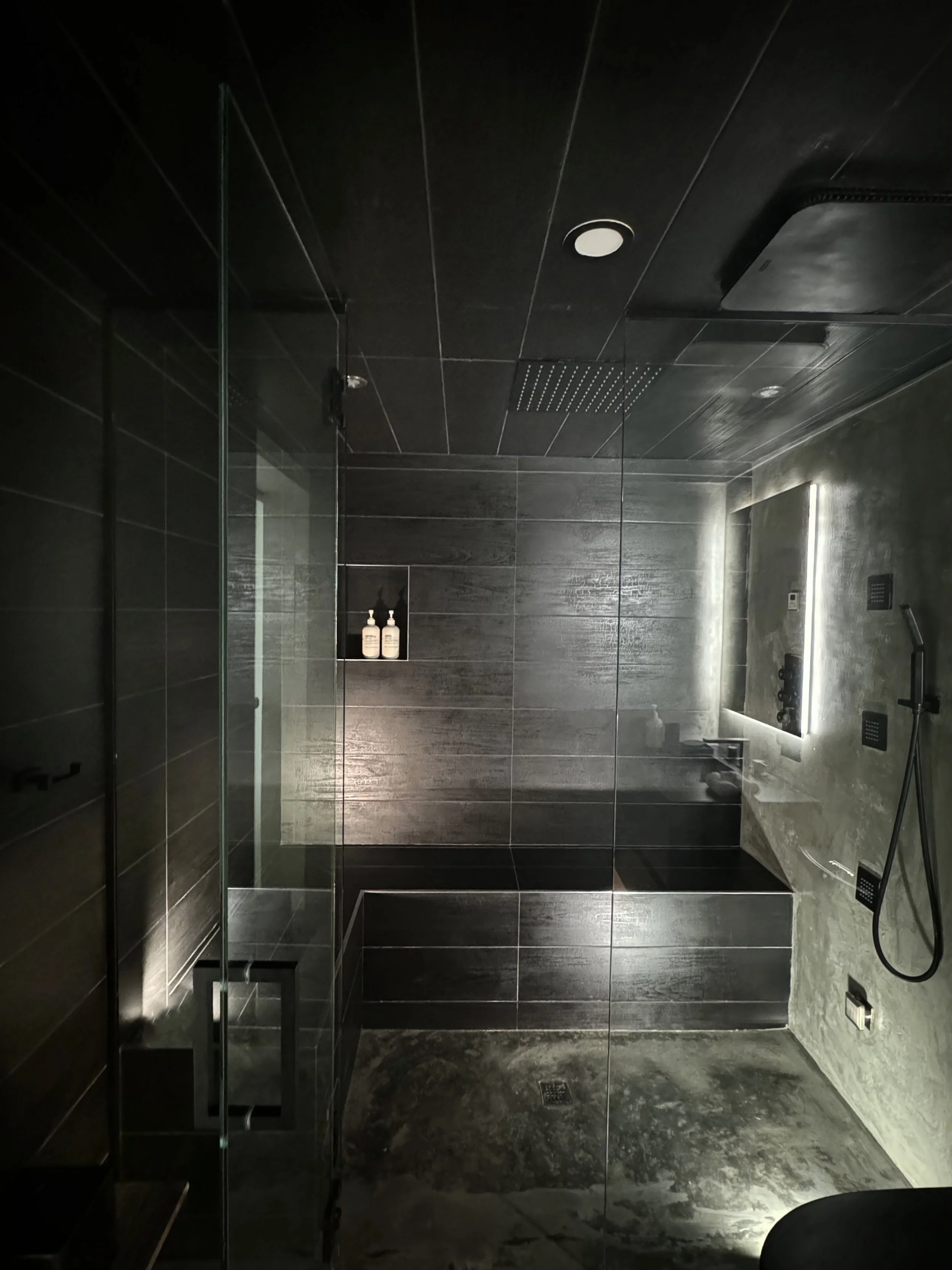 Modern, dark-toned bathroom with a glass shower enclosure, wall-mounted showerhead, lit mirror, and soap dispensers, featuring black tiles and minimalist design.