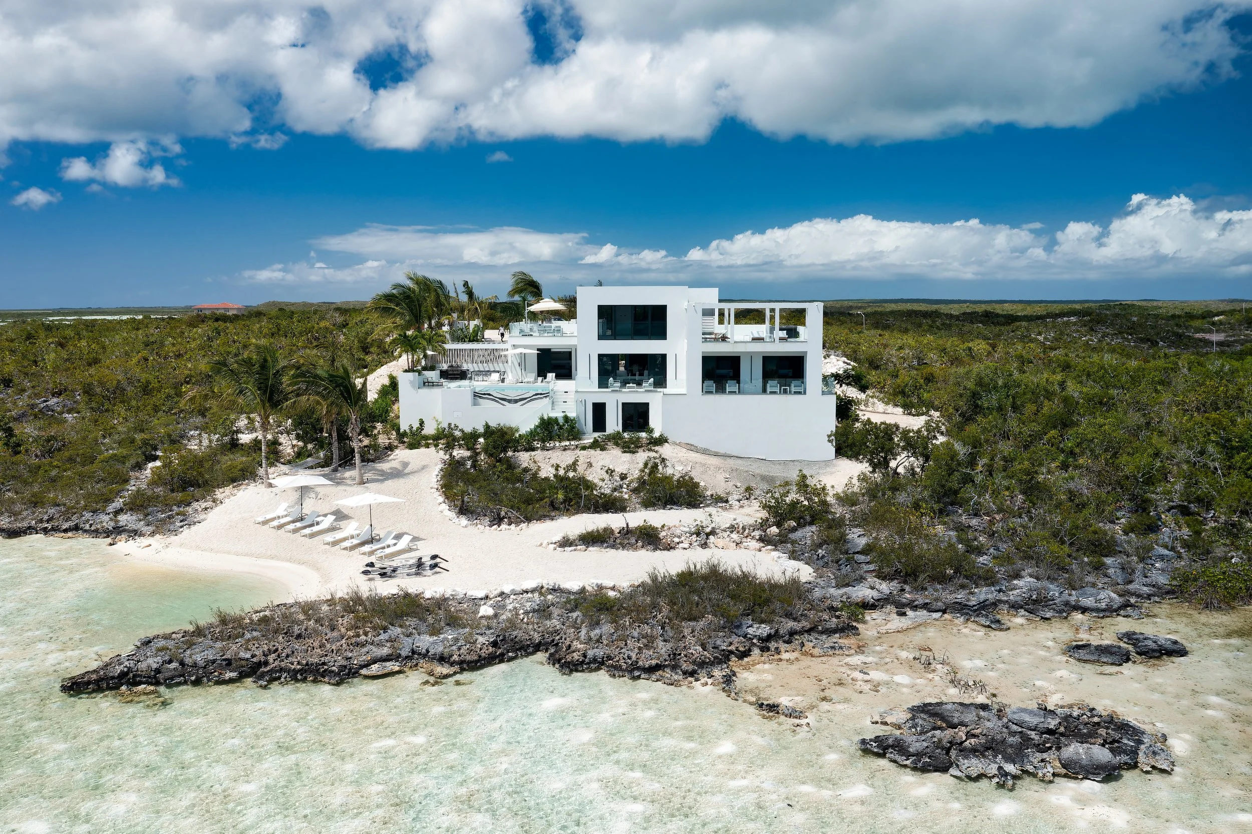 A modern white house situated on a sandy beach, surrounded by green bushes and palm trees, with lounge chairs and umbrellas near the shoreline under a partly cloudy sky.