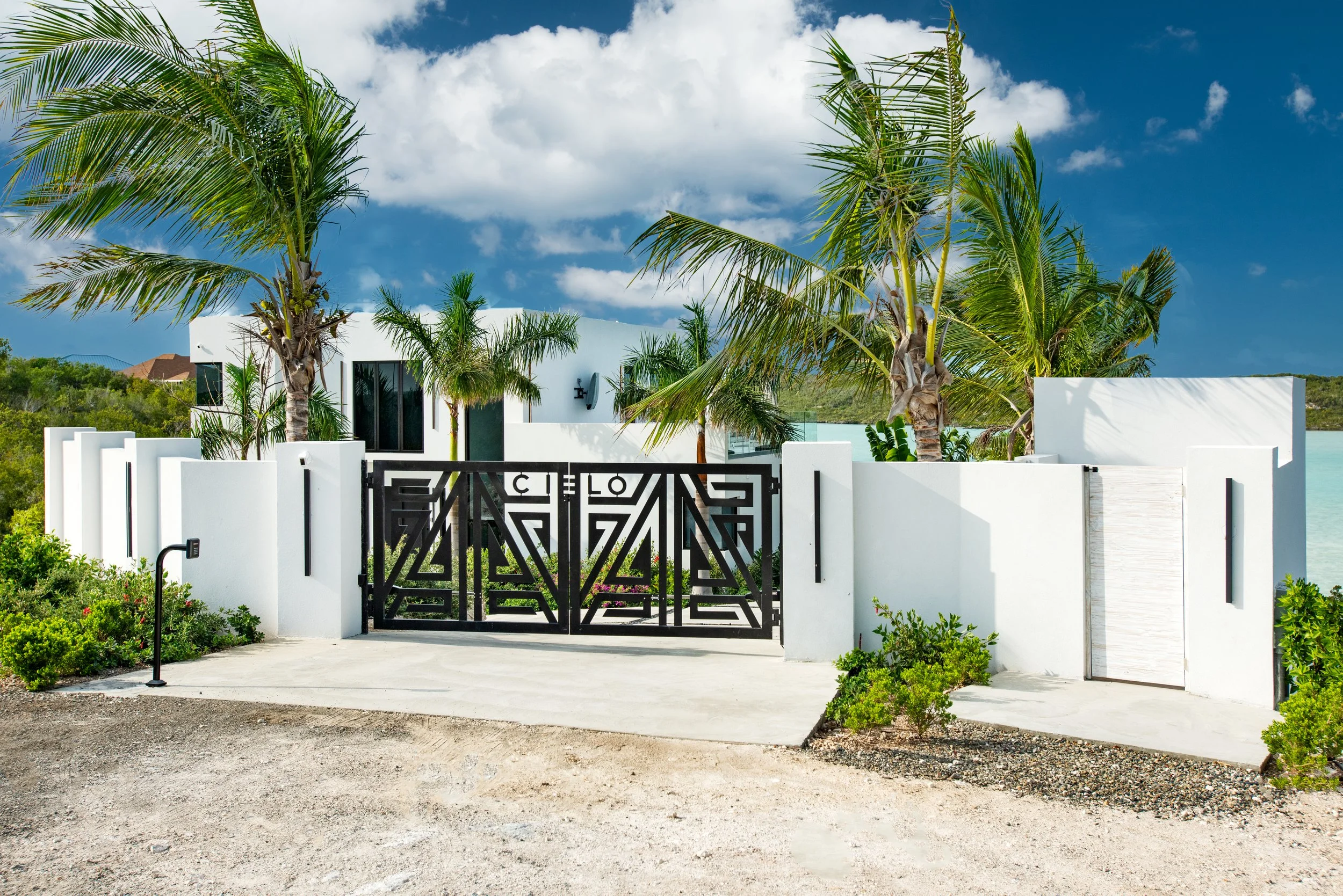 Modern white house with black gate surrounded by palm trees, near water, with a cloudy sky overhead.