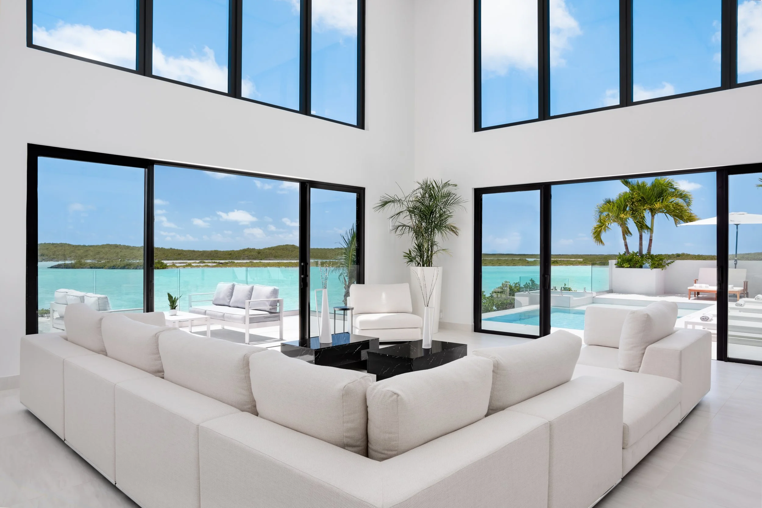 Modern living room with large windows overlooking a pool and ocean, white sectional sofa, black coffee tables, and potted plants.