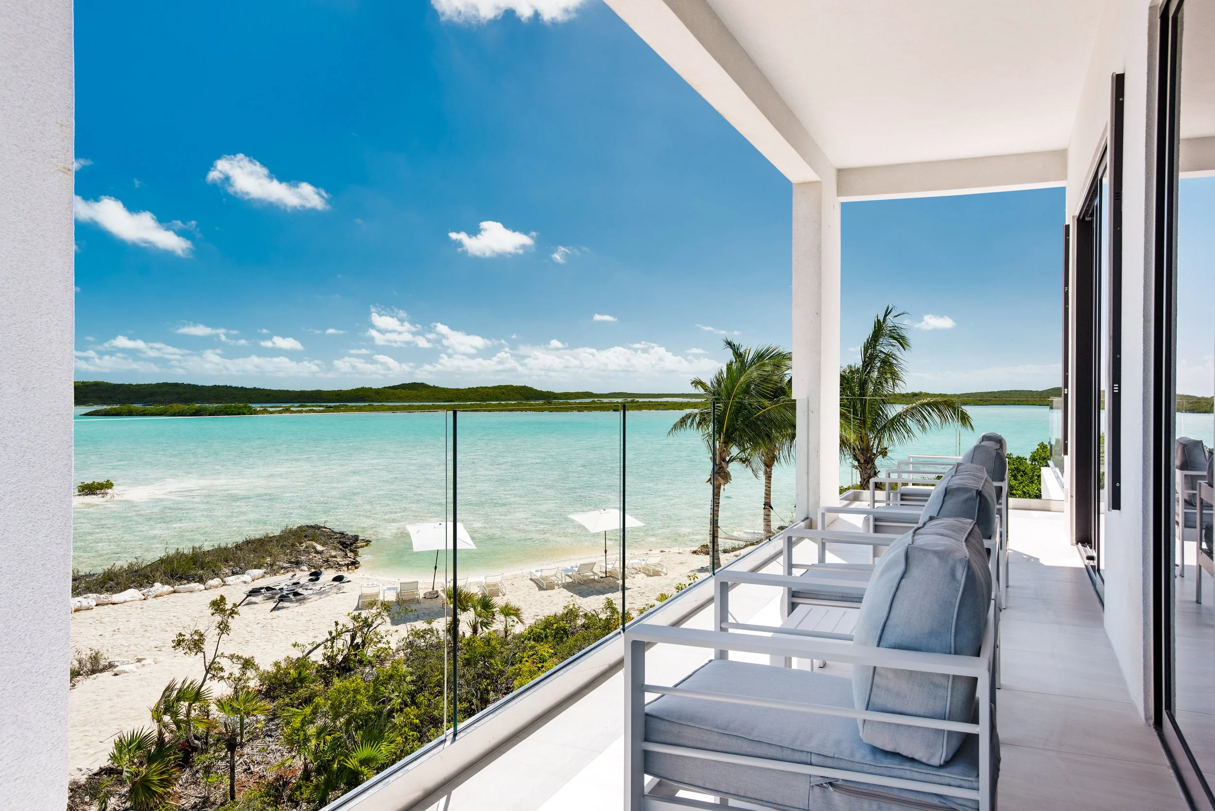Balcony overlooking a beach with lounge chairs, palm trees, and clear turquoise water under a blue sky with scattered clouds.