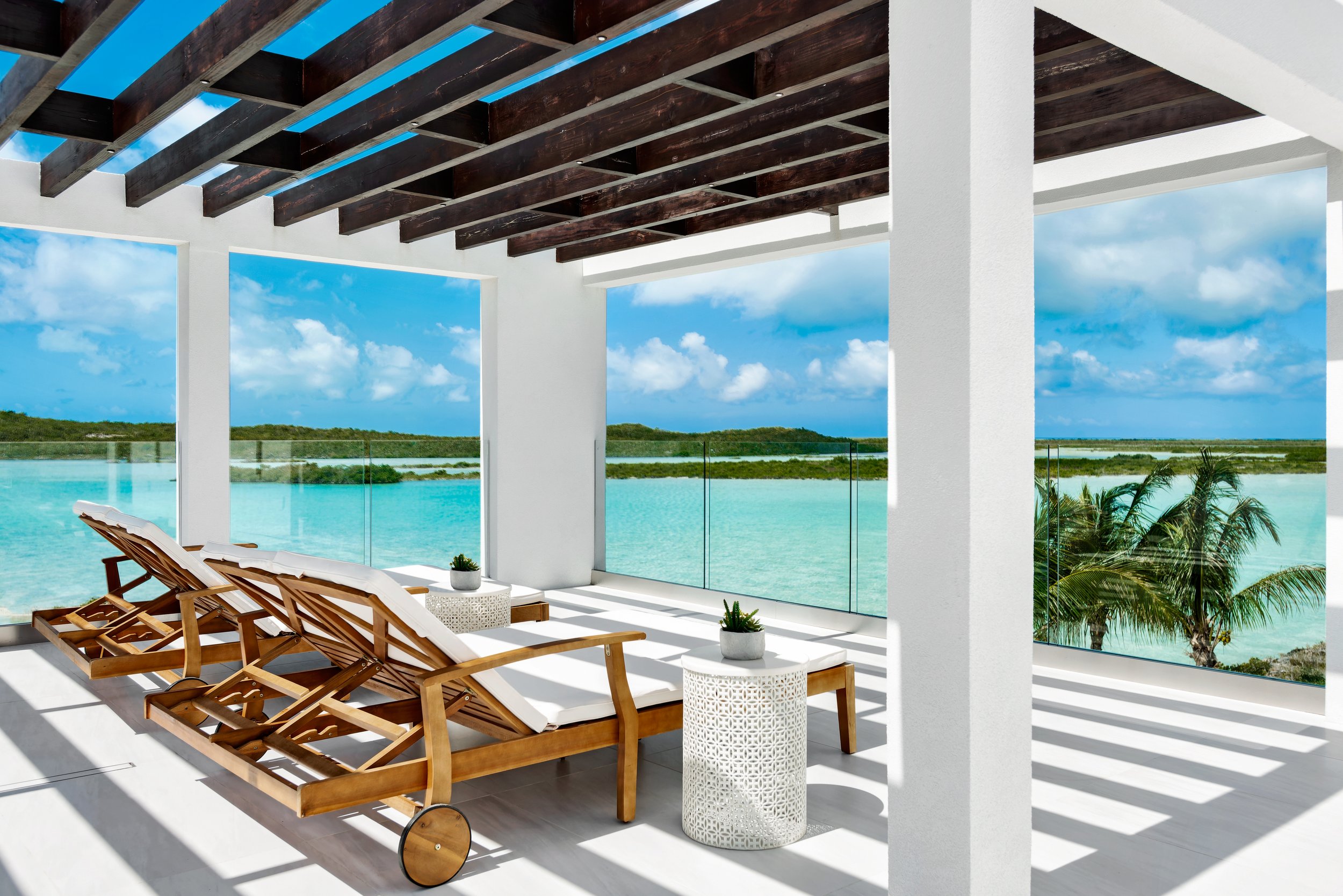 View from an open balcony overlooking a turquoise ocean with green islands, under a partly cloudy sky. The balcony has white walls, dark wooden beams on the ceiling, wooden lounge chairs with white cushions, and white side tables with small potted pl