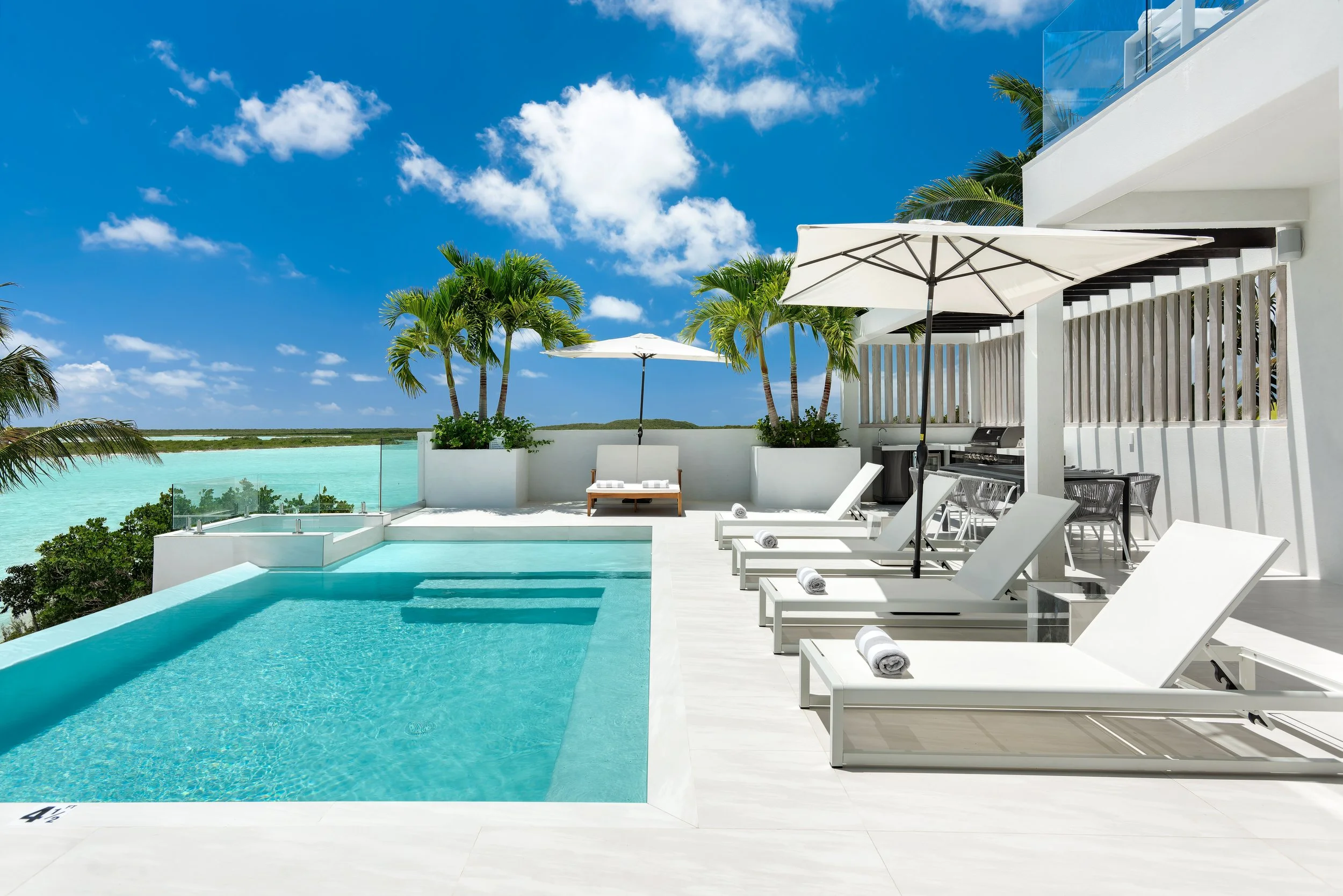 Luxury poolside with white lounge chairs, umbrellas, and palm trees, overlooking a turquoise water landscape under a blue sky with clouds.