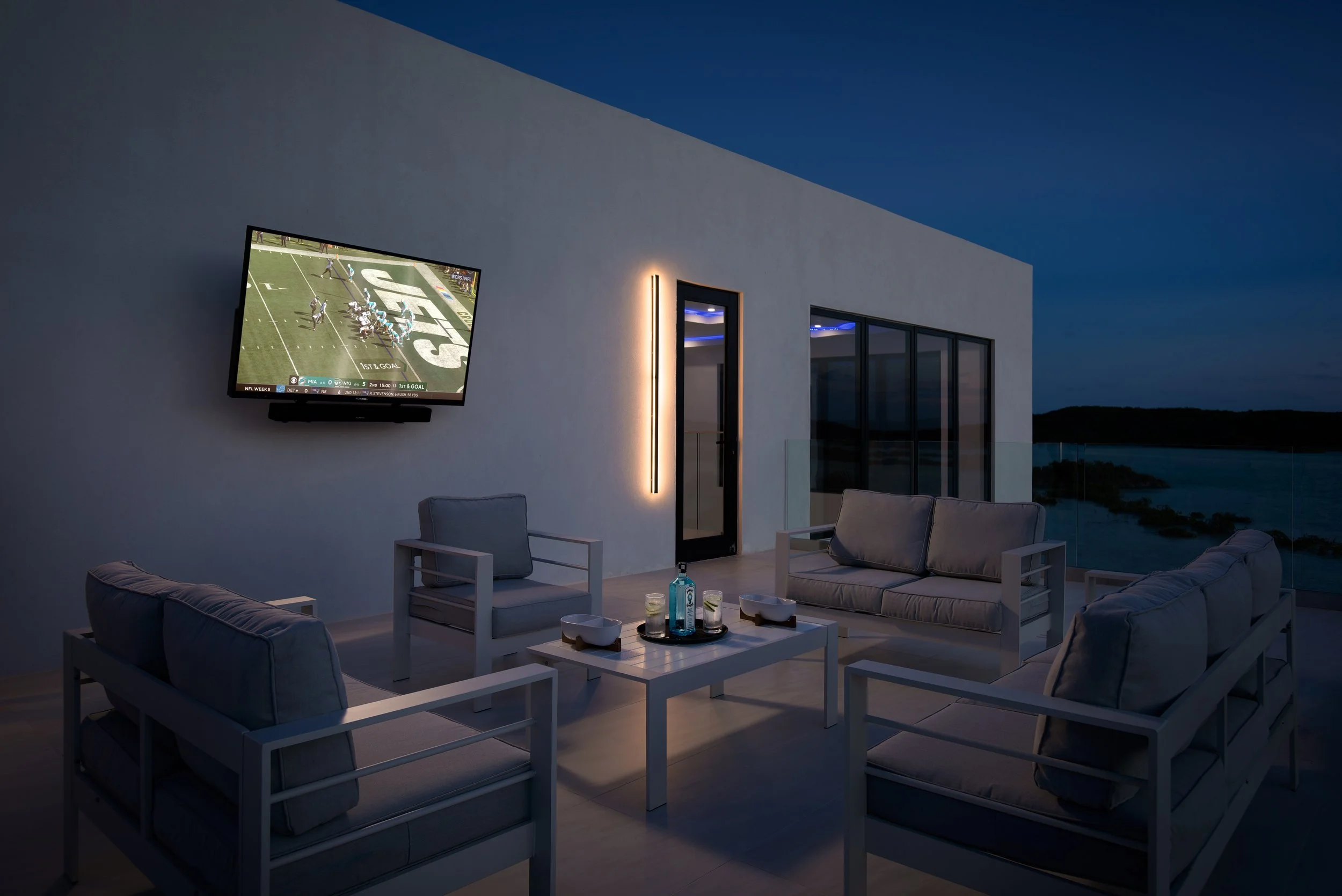 Outdoor terrace at dusk with a mounted TV showing a football game, white outdoor furniture, a small table with drinks, and between the terrace and sky are glass railings with a water view.