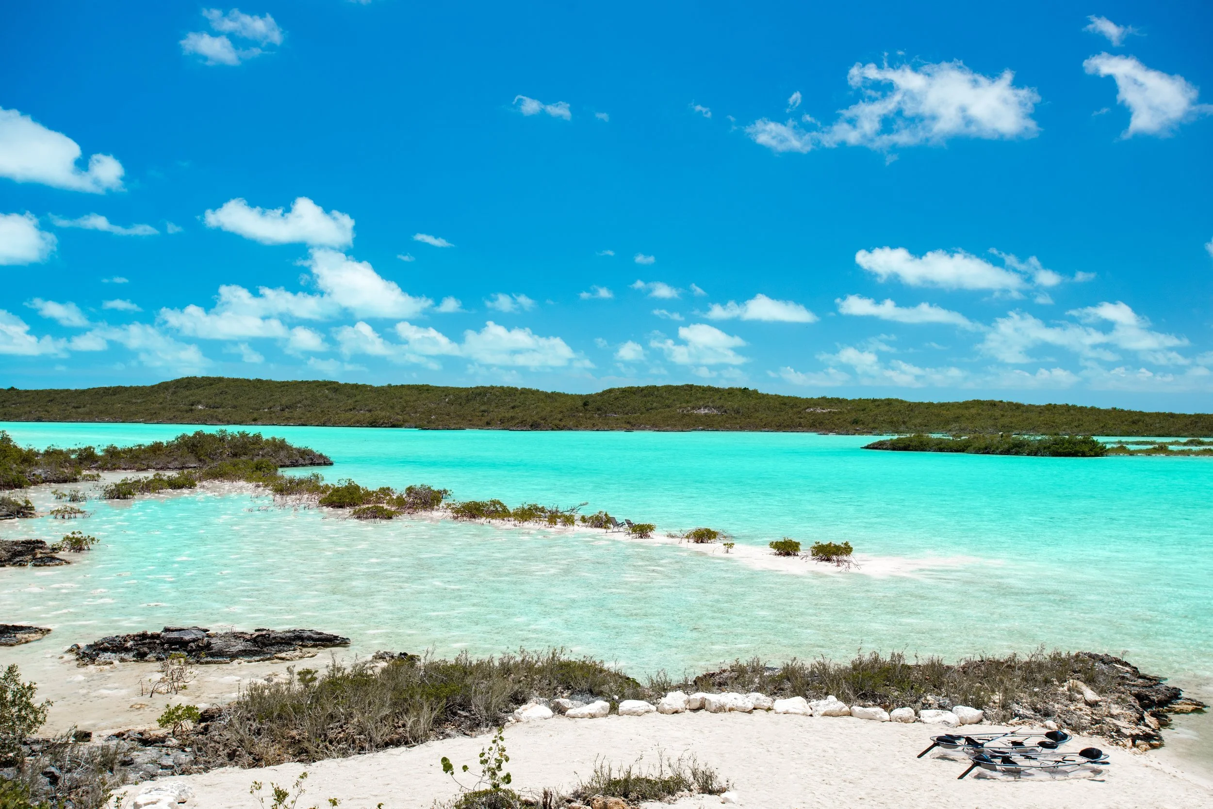 Tropical beach with white sand, turquoise water, green shrubbery, and a blue sky with scattered clouds.