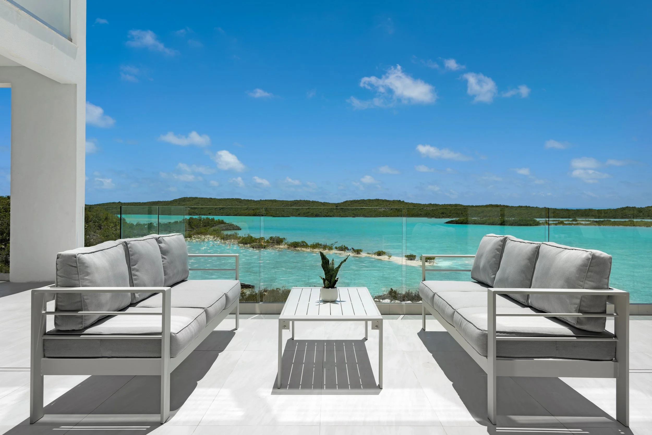 Photo of a balcony with two white sofas and a white coffee table with a potted plant, overlooking a turquoise lake with small islands and a partly cloudy blue sky.