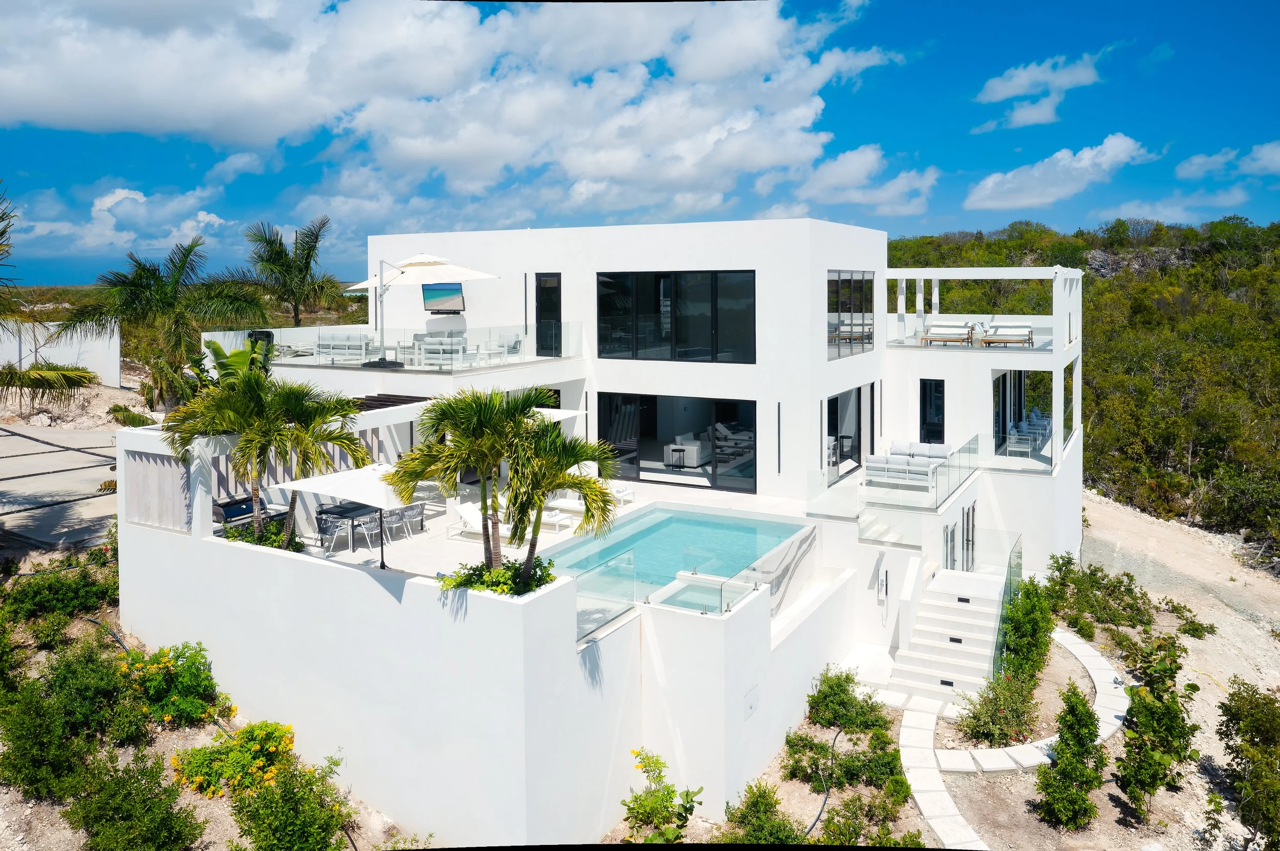 Modern white multi-story house with outdoor pool, patio furniture, and palm trees, situated on a lush landscape under a bright blue sky with scattered clouds.