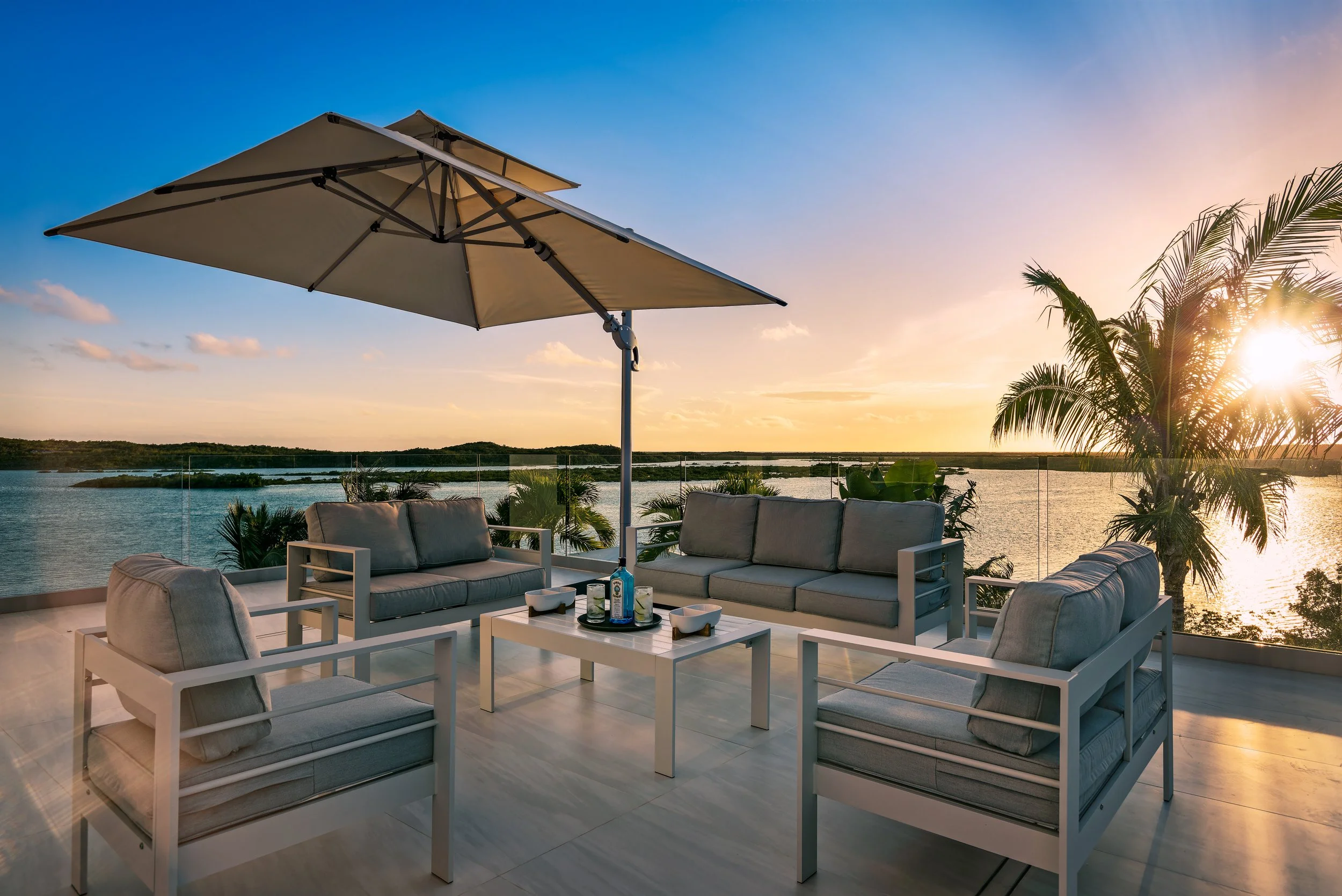 Outdoor patio with white furniture and a large beige umbrella overlooking a body of water at sunset, with trees and a clear sky.