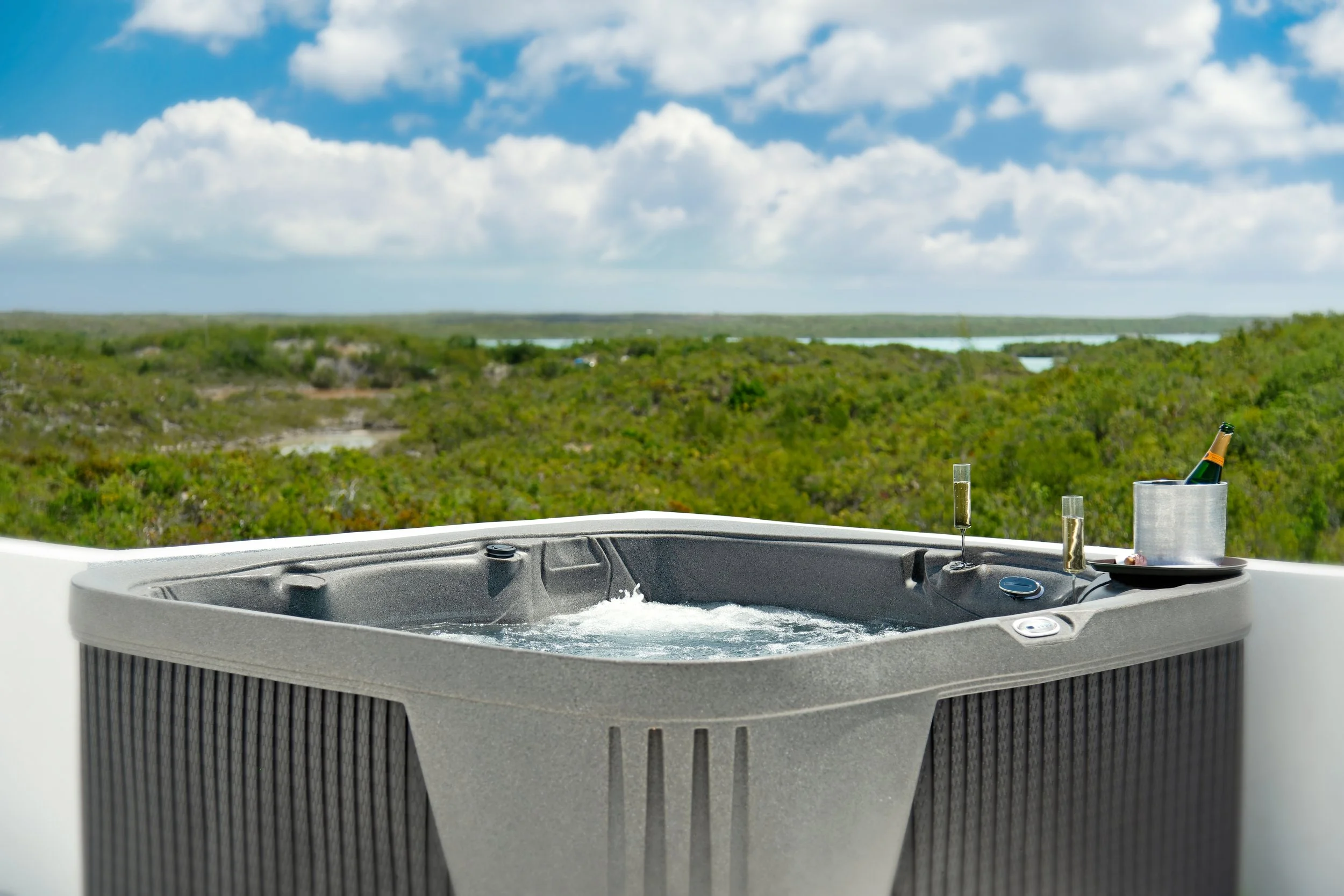 A hot tub on a balcony with a scenic view of green vegetation, lakes, and a blue sky with clouds, with a bottle of wine and glasses on a tray.