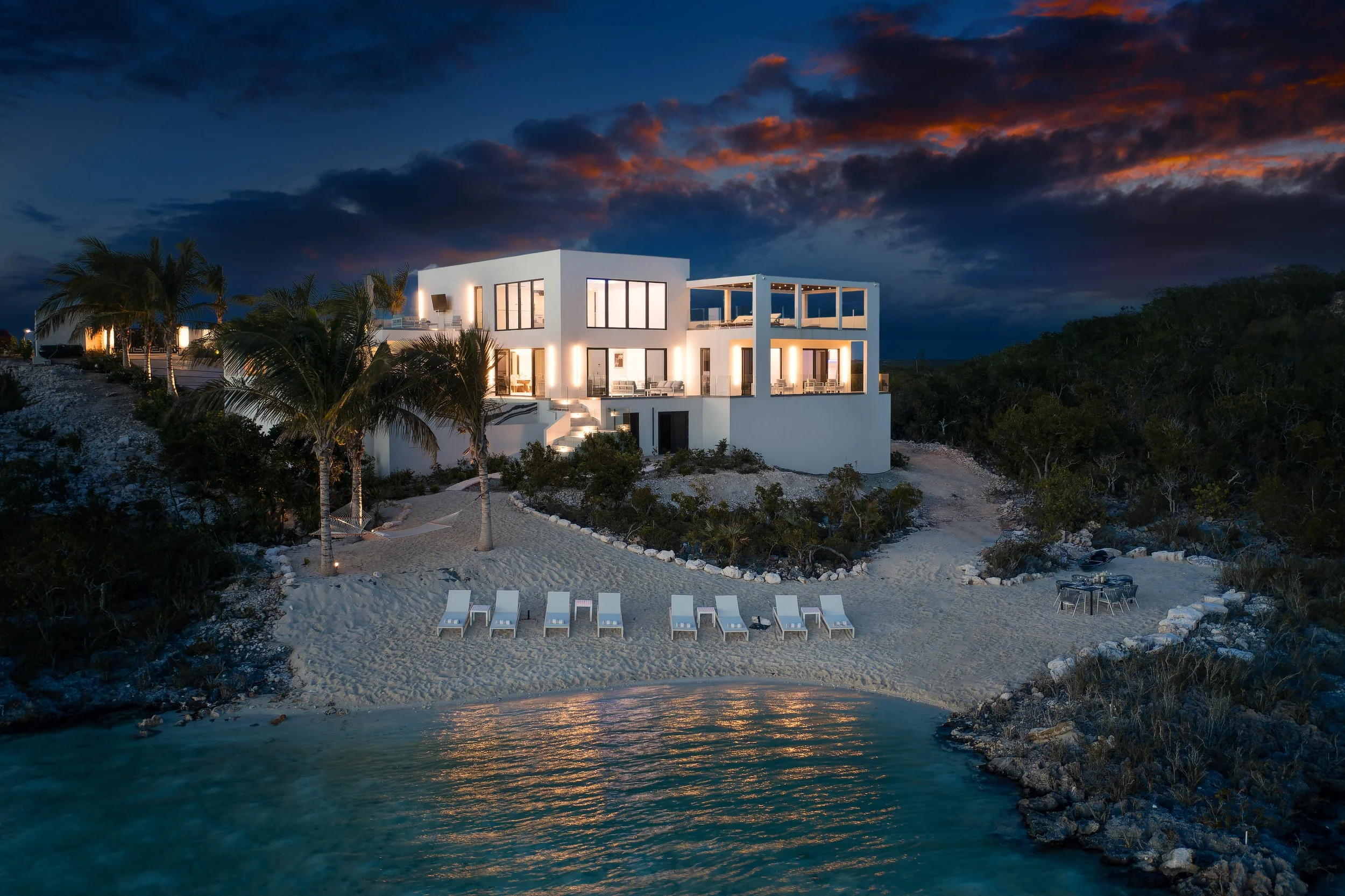 A modern white house on a beach at dusk with illuminated interior lights, palm trees, sun loungers on the sand, a small table and chairs, and a calm water body in the foreground.
