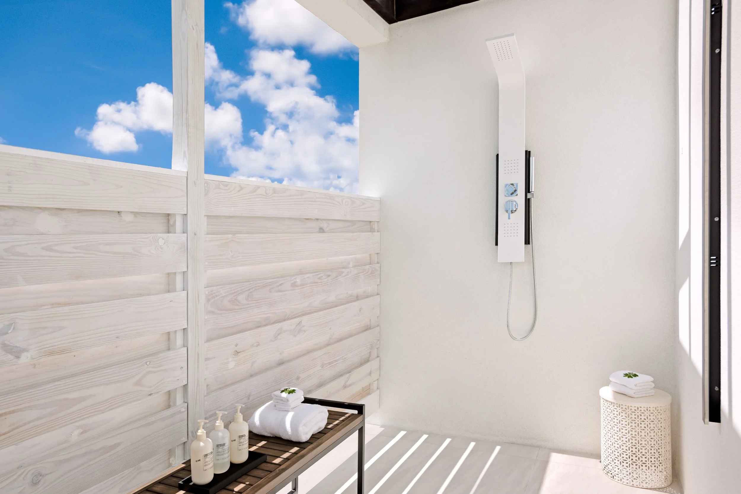 Outdoor shower area with white walls, a large white shower panel, and a small table holding towels and lotions. The space is open to a sky with clouds, and the floor has white slats allowing sunlight to cast striped shadows.