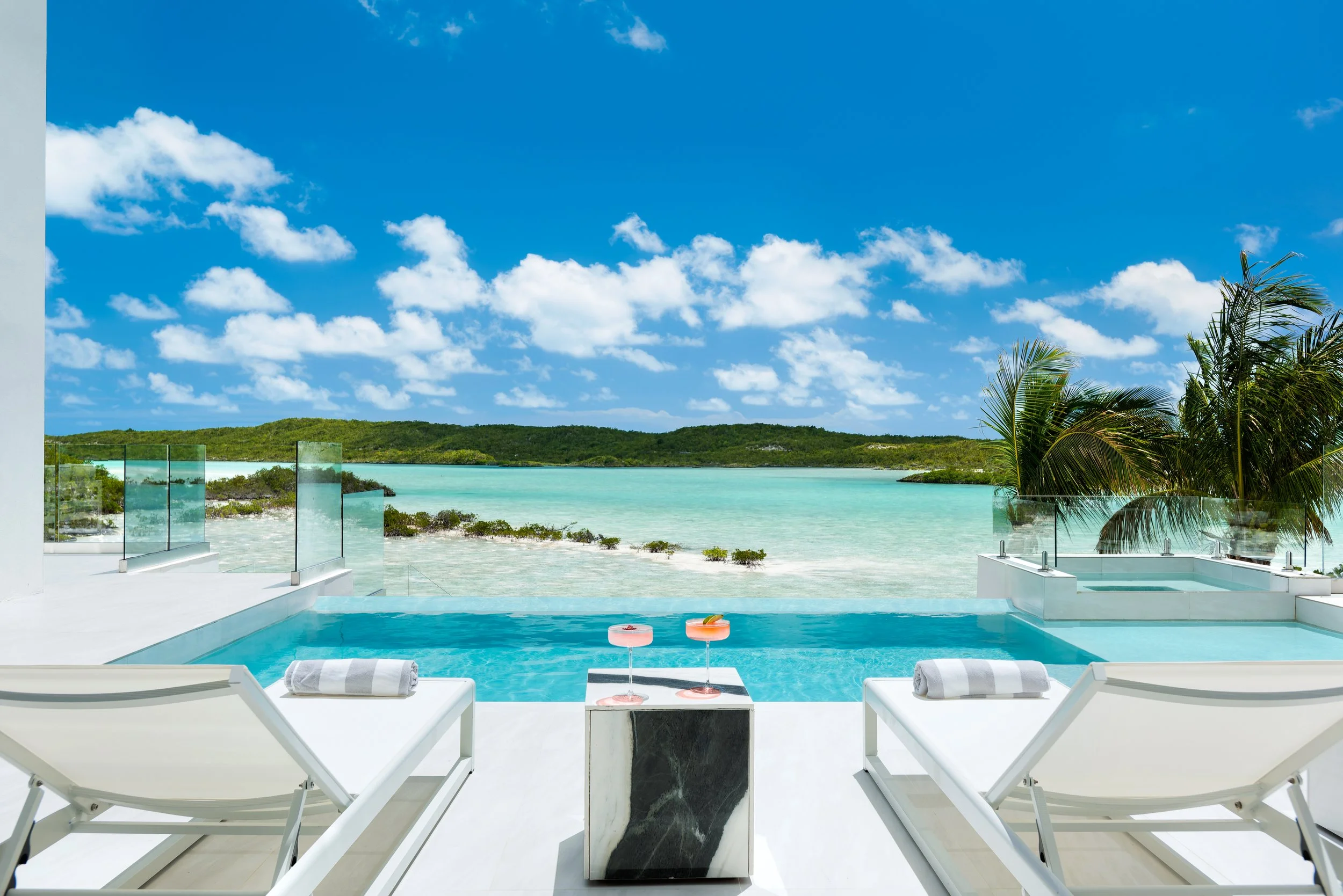 A luxurious outdoor pool area overlooking a clear blue ocean with a green island in the distance on a sunny day with a partly cloudy sky.
