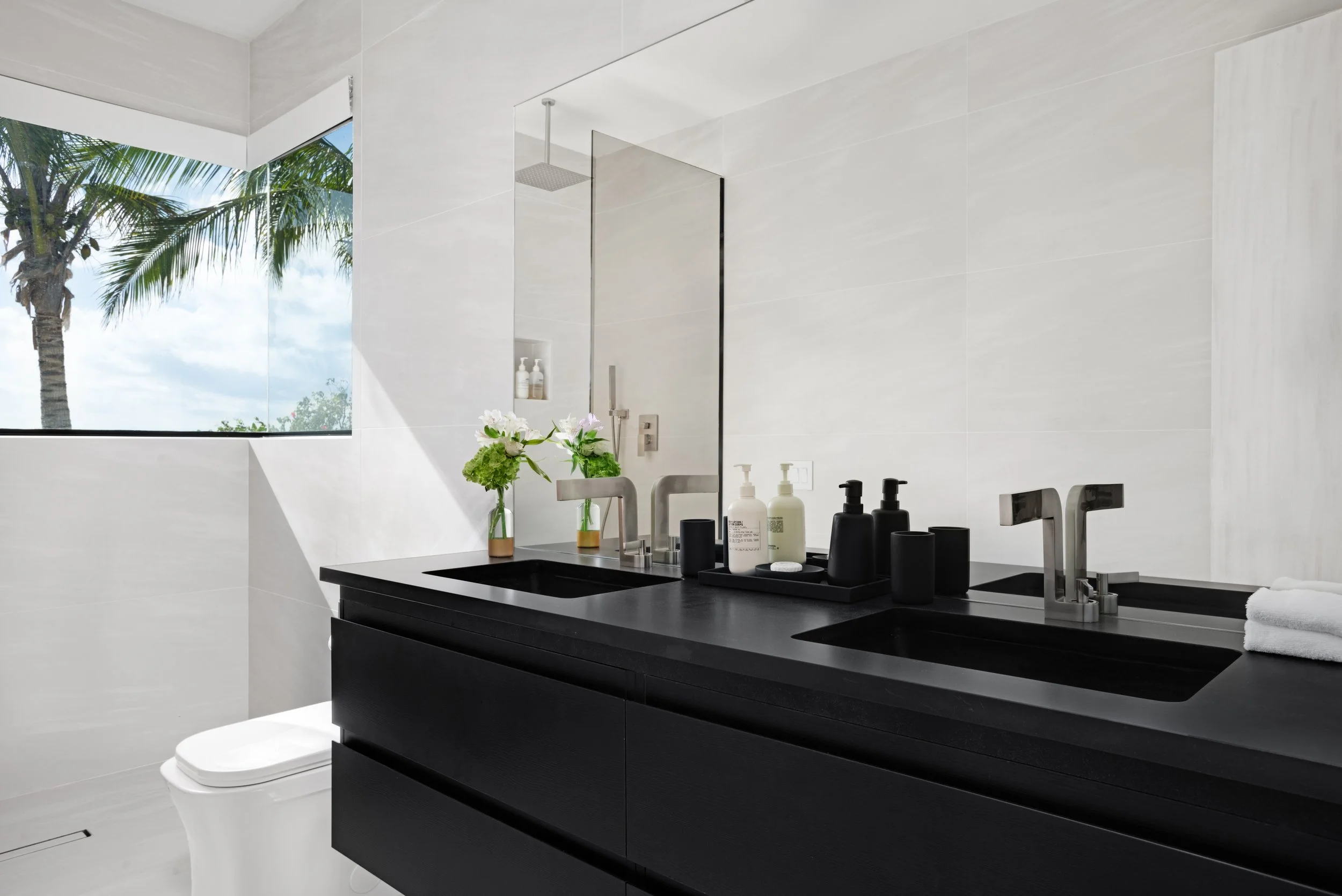 Modern bathroom with black double sinks, large mirror, white walls, a window showing palm trees outside, and decorative vases with flowers on the counter.