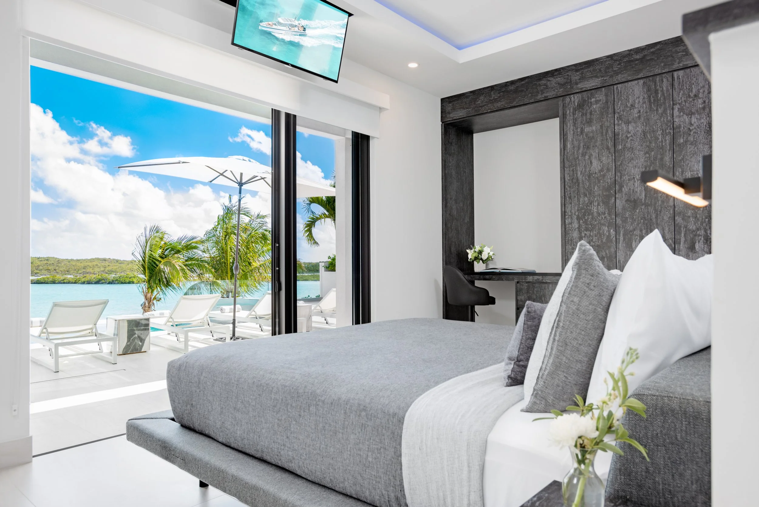 Modern bedroom with a large bed, gray bedding, and white pillows, overlooking a balcony with lounge chairs, umbrellas, palm trees, and a water body under a blue sky with clouds.