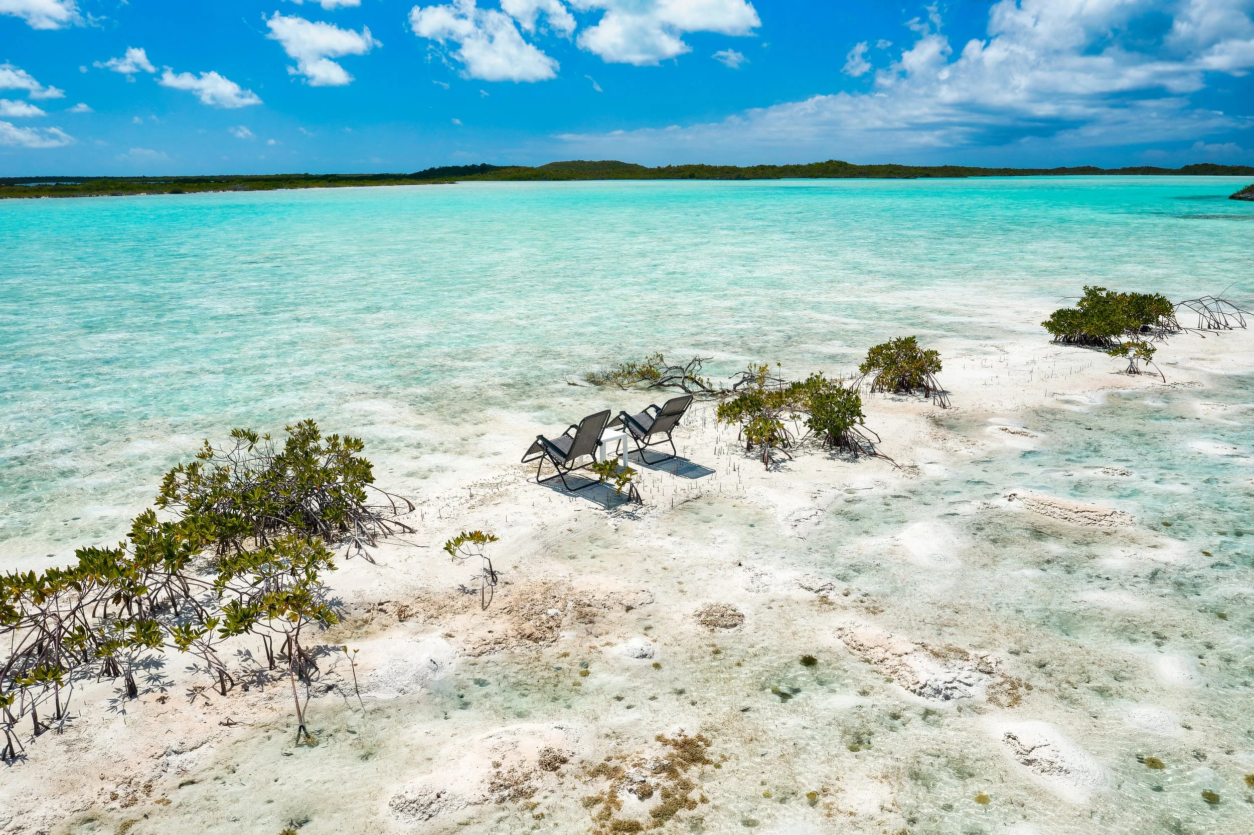 Two lounge chairs on a sandy beach with small bushes, overlooking clear turquoise water and a distant green landmass under a blue sky with scattered white clouds.