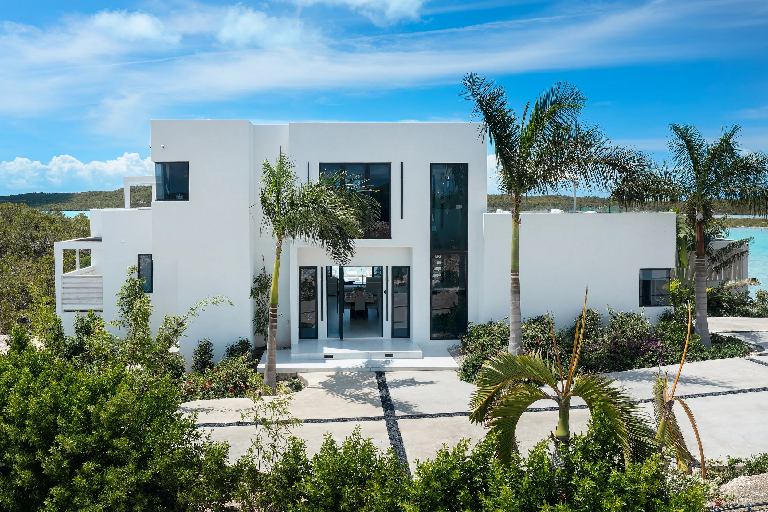Modern white house with palm trees in front, surrounded by greenery, under a blue sky with clouds.