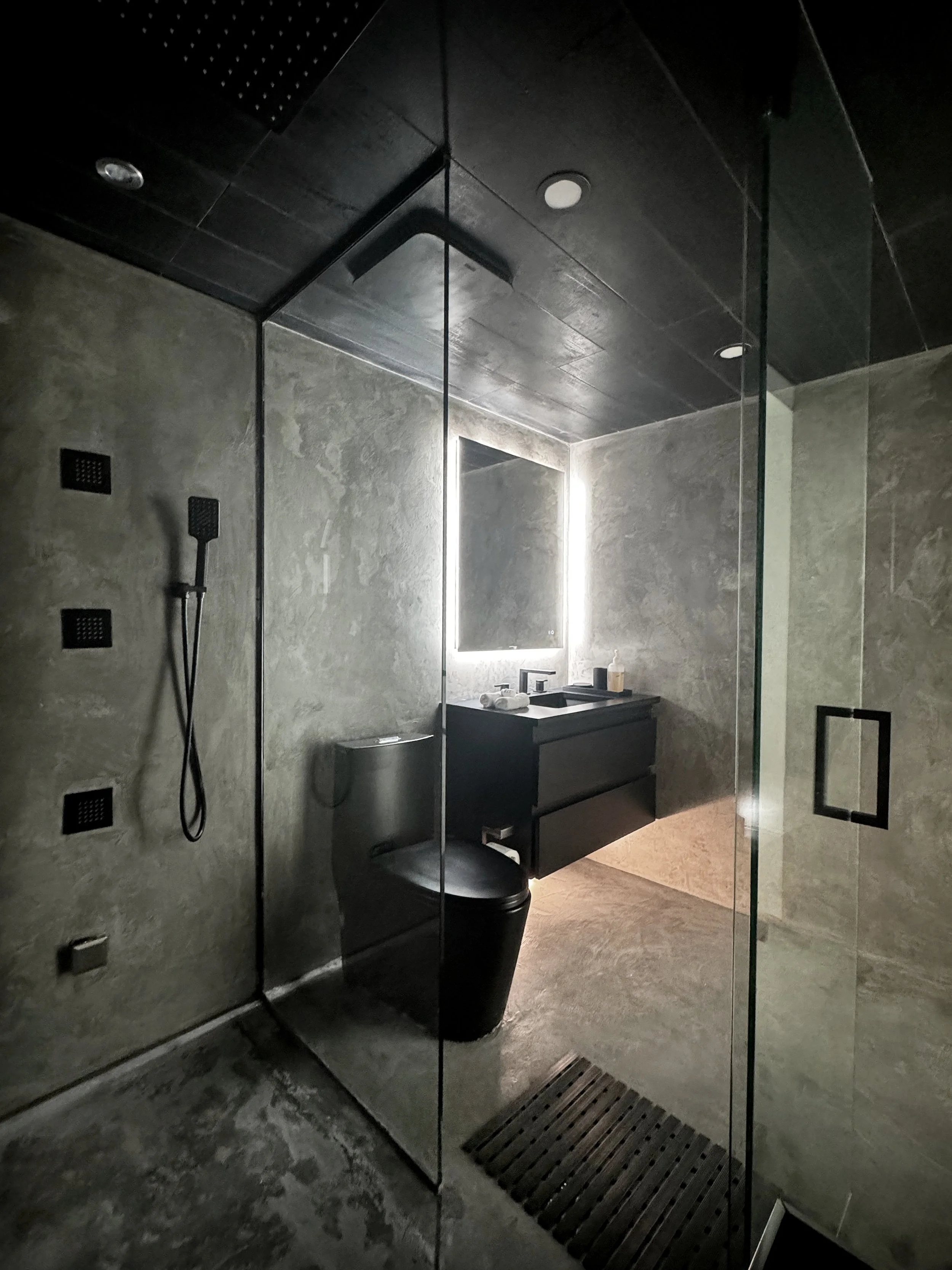 Modern bathroom with concrete walls and floor, black toilet, and black vanity with illuminated mirror. Glass shower enclosure with black handle and showerheads.