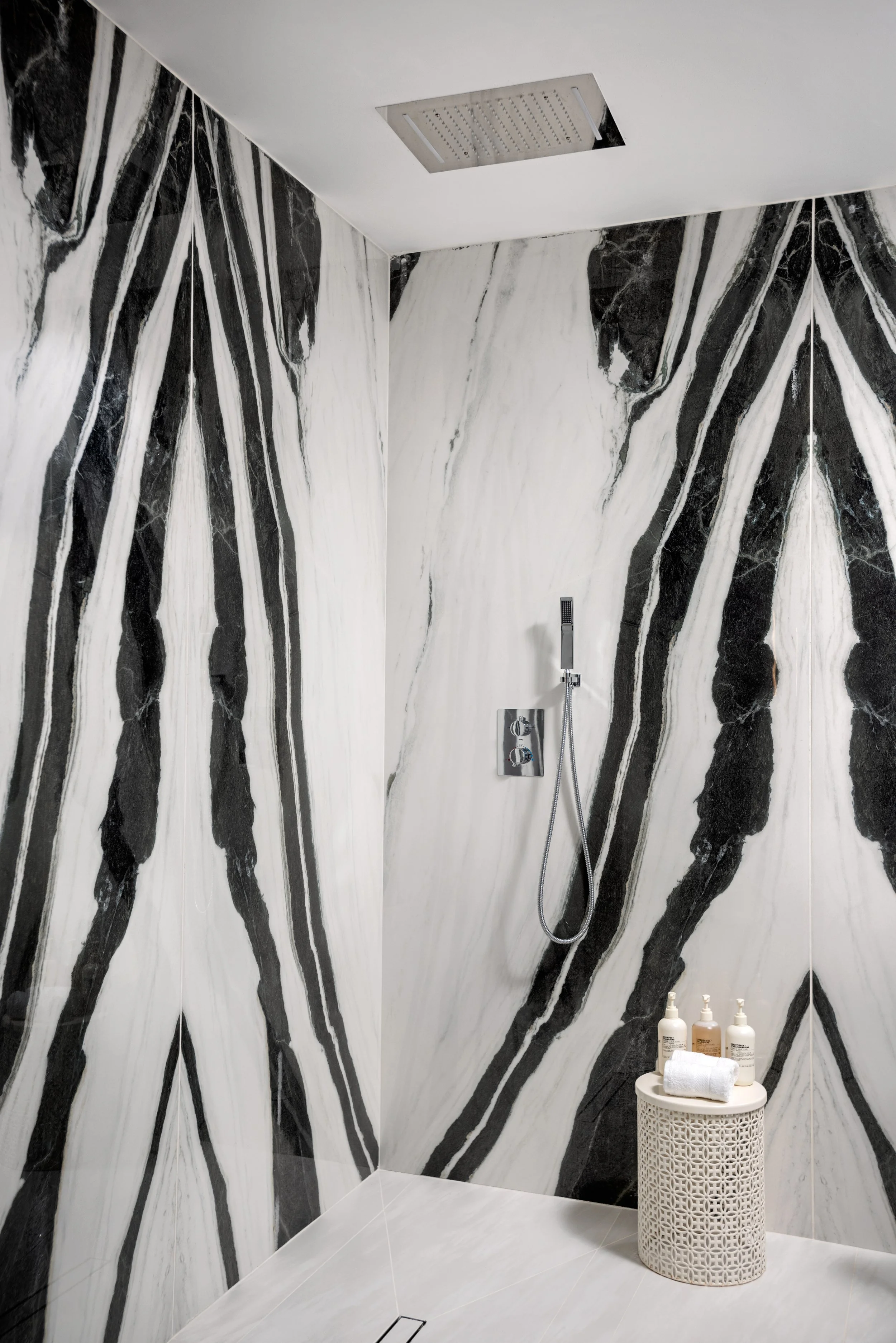 A modern shower with black and white marble tiles on the walls, a ceiling-mounted showerhead, a handheld shower on a wall panel, and a small round table with bottles of soap and a towel.