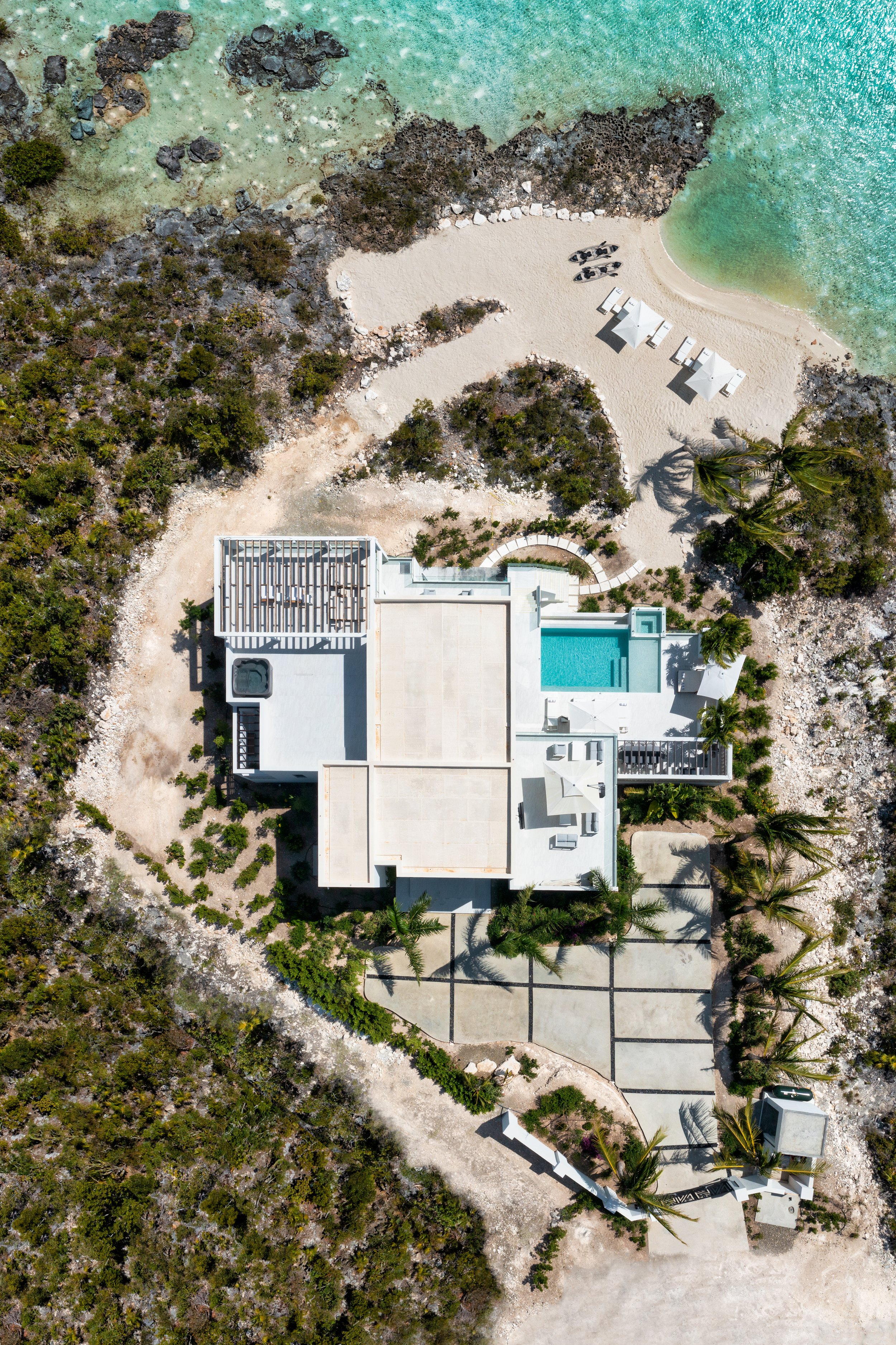 Aerial view of a modern beachfront house with a swimming pool, surrounded by lush greenery and rocky terrain, with a sandy beach and turquoise ocean nearby.