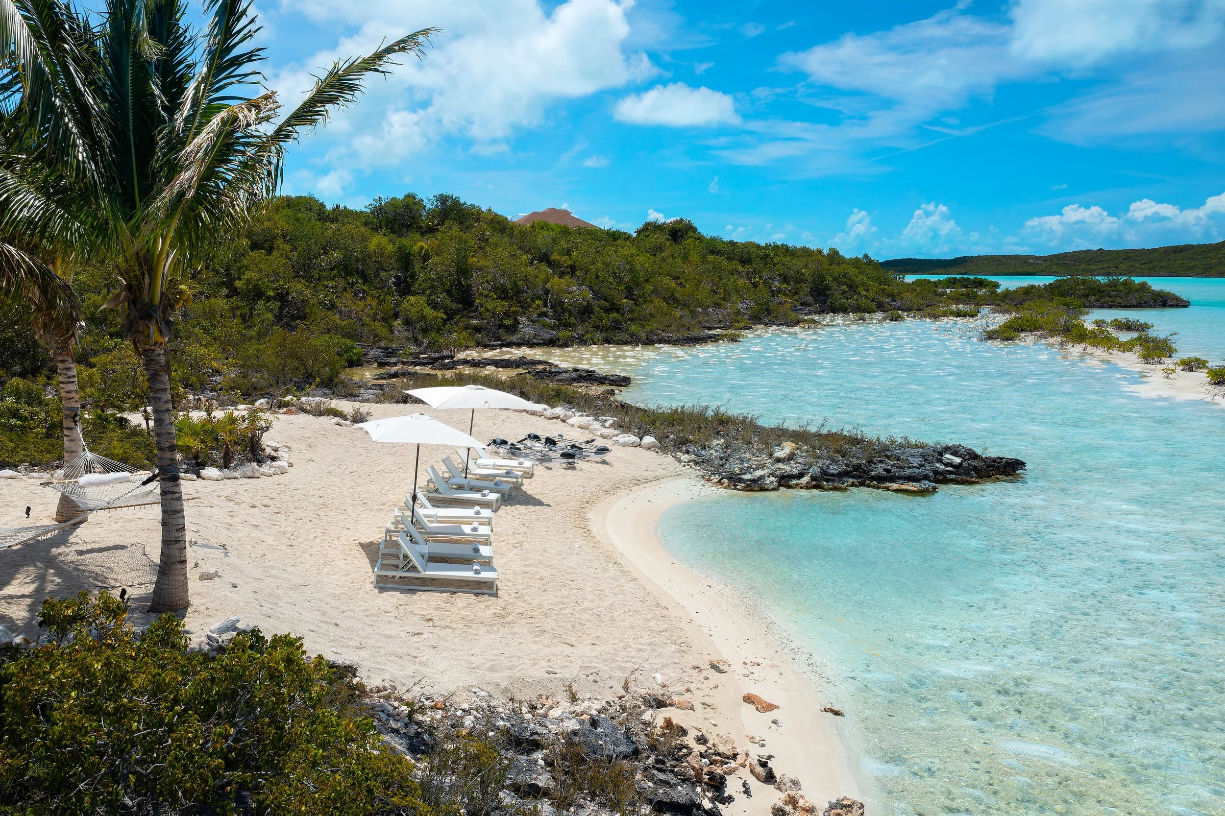 A tropical beach scene with white sand, turquoise water, green vegetation, palm trees, and a row of white lounge chairs with umbrellas.