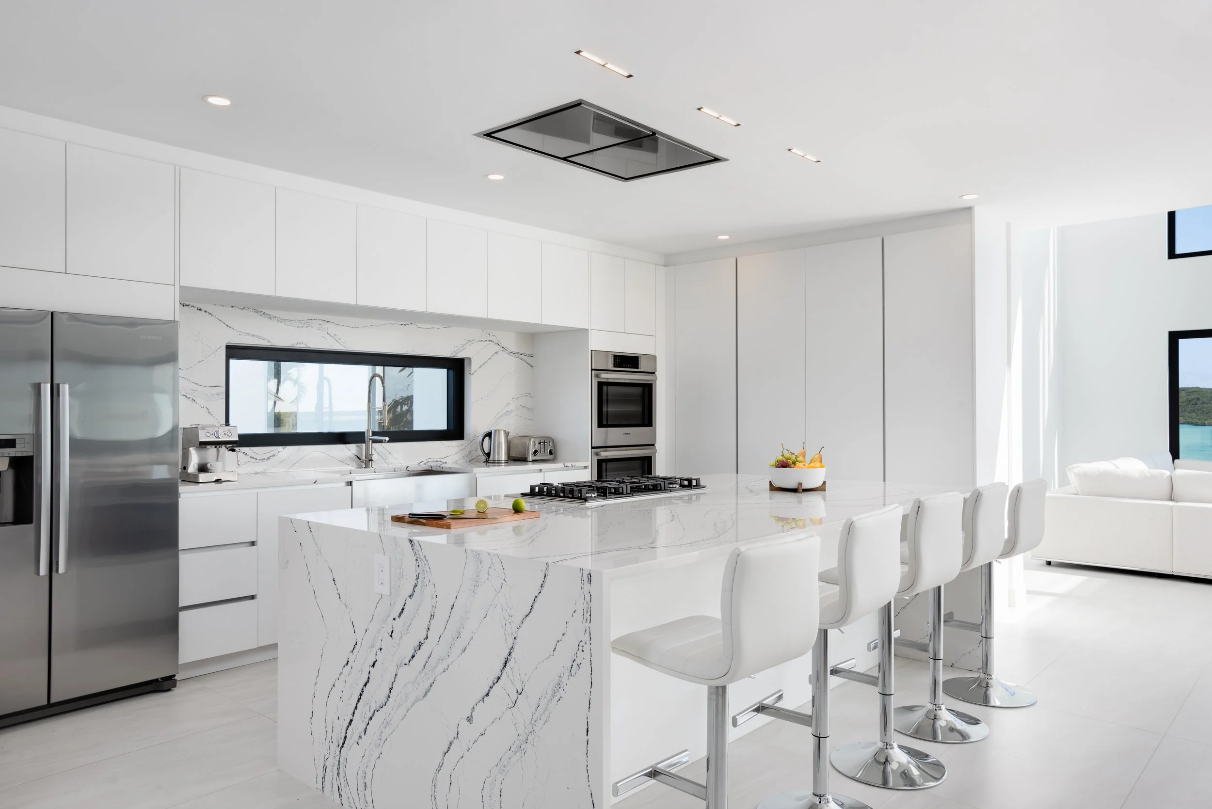 Modern kitchen with white cabinetry, marble island, stainless steel appliances, bar stools, and ocean view through windows.