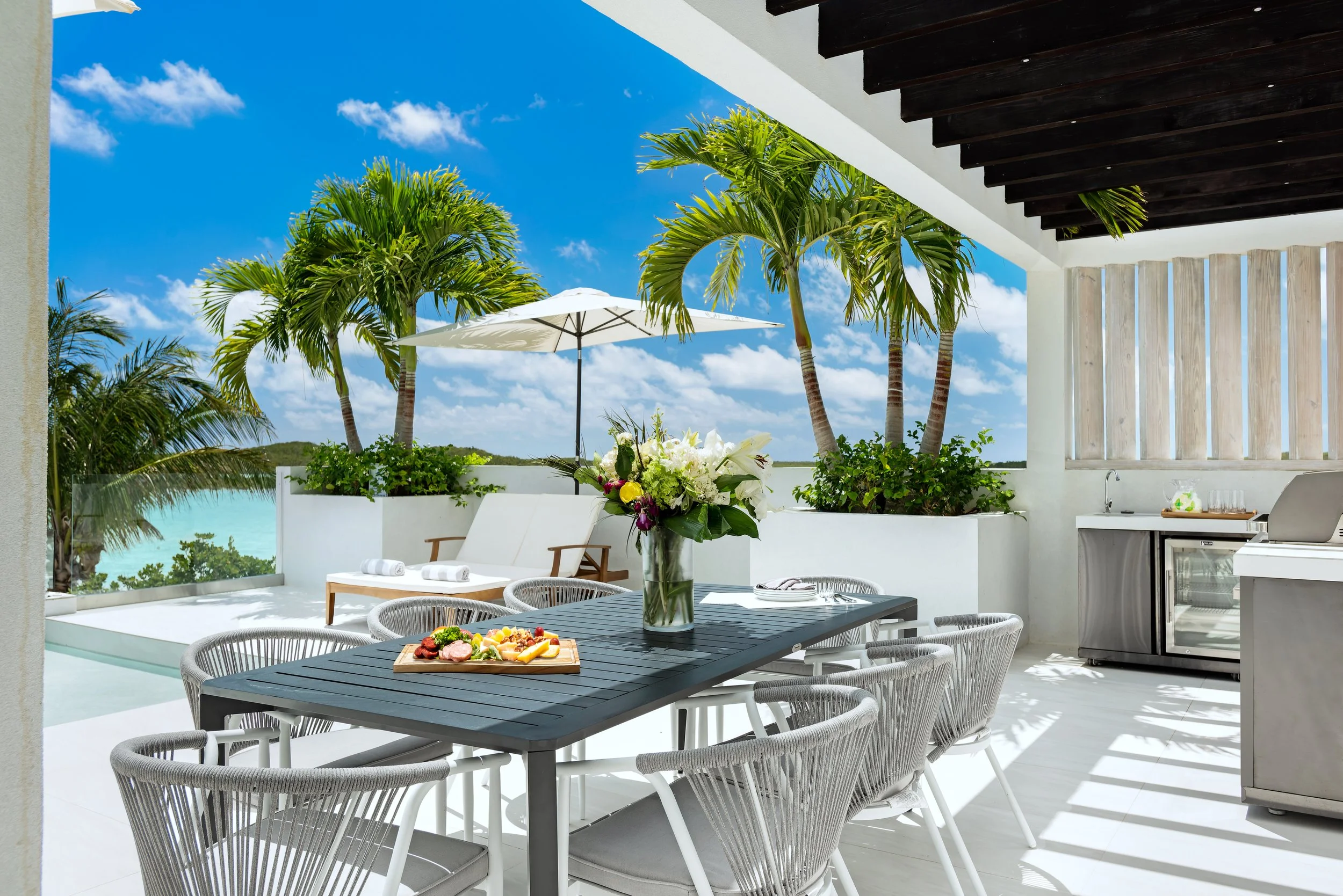 Outdoor patio with a dining table, surrounded by chairs, a vase of flowers, an outdoor kitchen, palm trees, and a view of the ocean under a blue sky with clouds.