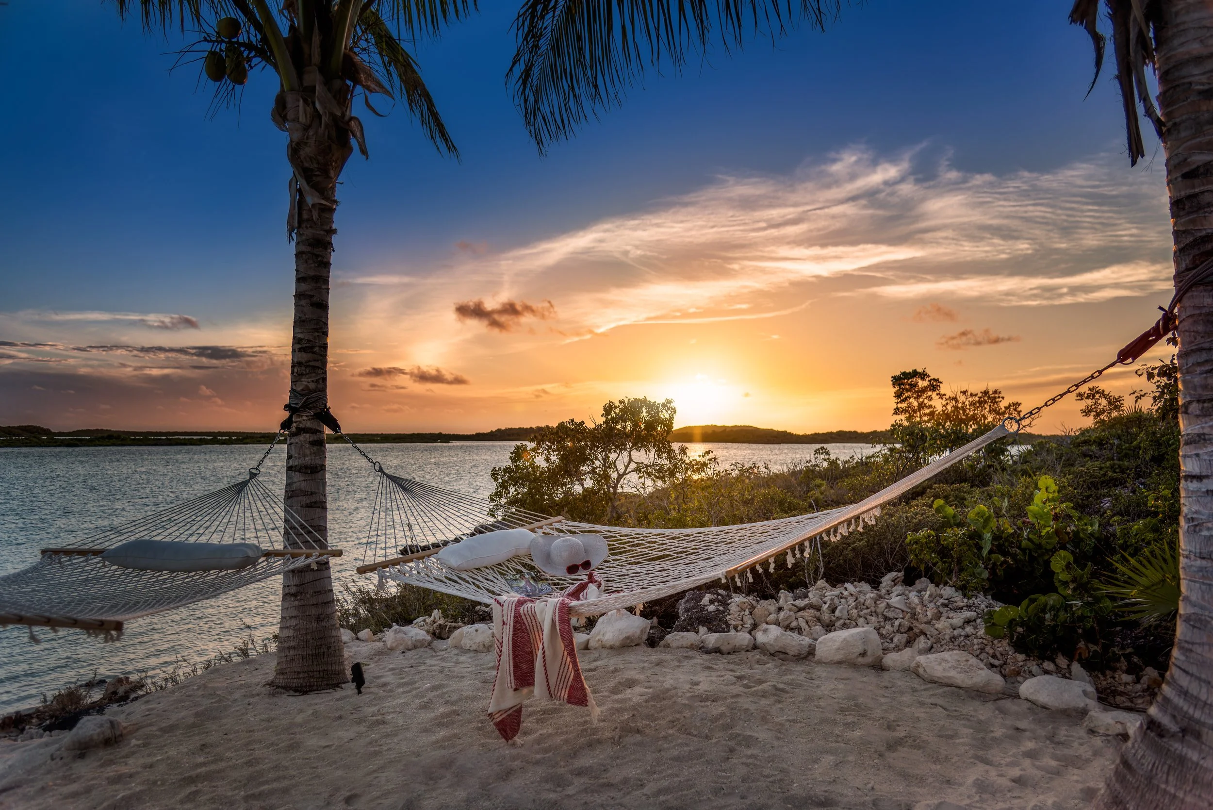 Hammock strung between two palm trees on a sandy beach by a body of water during sunset, with a towel and sunglasses hanging on the hammock.