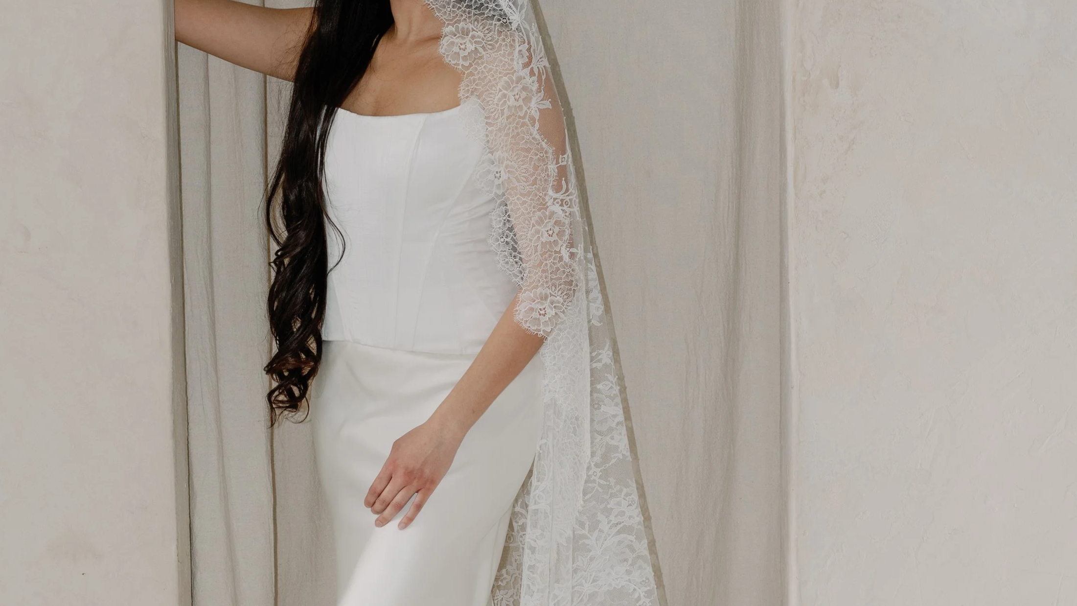 Close-up of a woman wearing a white strapless dress with lace details on the shoulder and a lace veil, standing against a beige wall.
