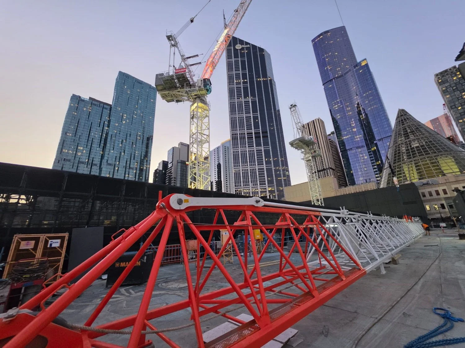 Red and white tower crane jib laid out on site at dusk below Melbourne CBD towers