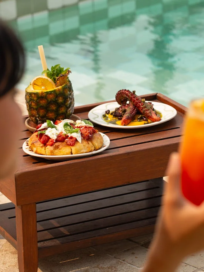 A wooden table set with a variety of dishes including grilled steak with mustard, grilled prawns with lemon, grilled corn, green vegetable salad, dark red wine, and small sandwiches with toppings, with a pineapple-shaped container holding drinks.