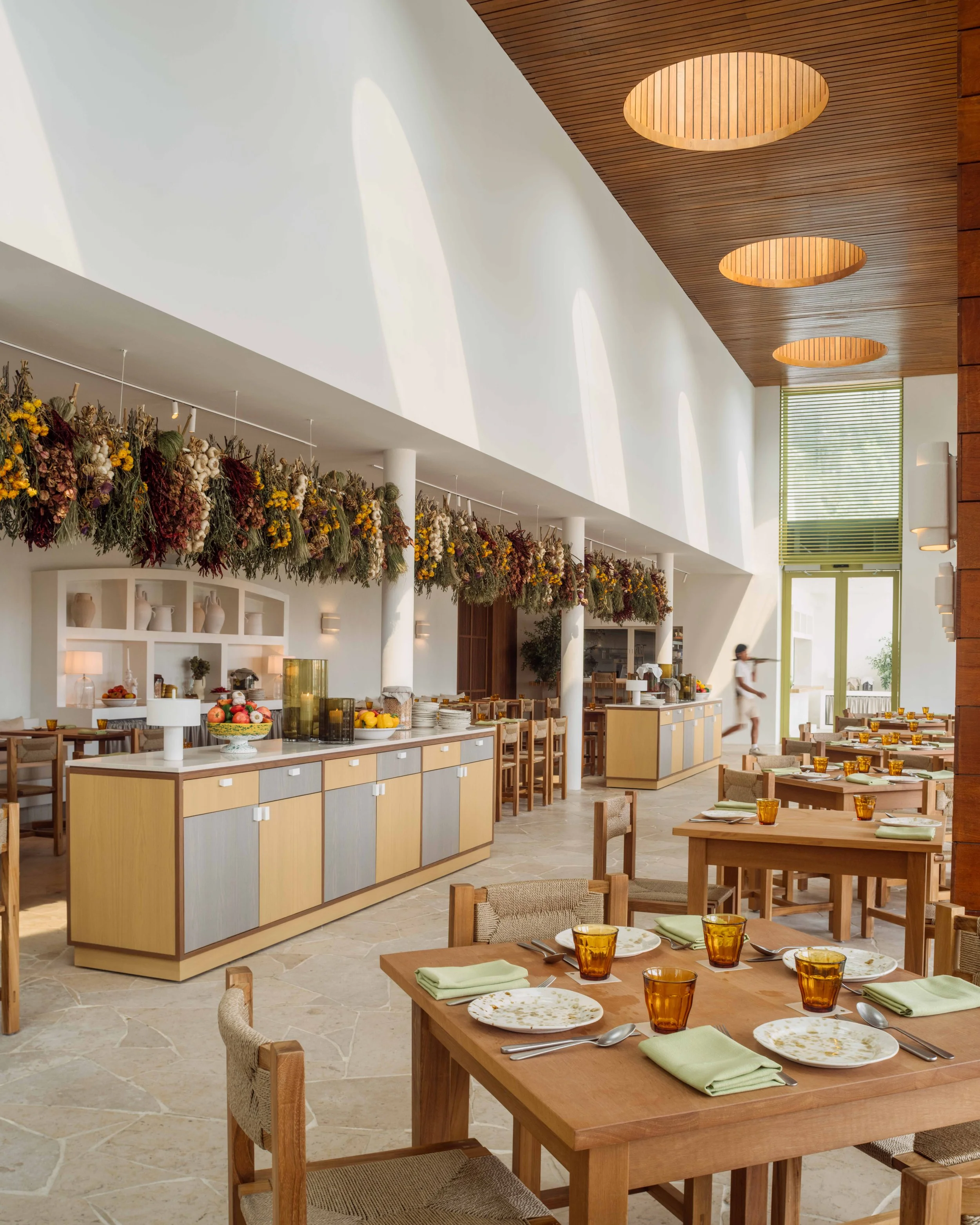 Empty restaurant with wooden tables and chairs, marble floor, large windows, ceiling fans, and greenery on the upper level.