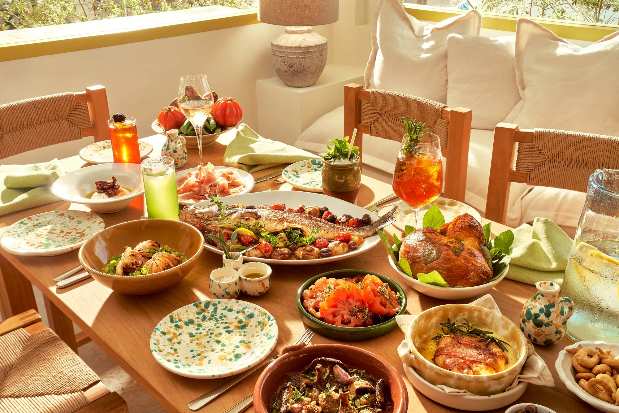 Empty restaurant with wooden tables and chairs, glass holders, and modern decor including a ceiling fan, plants, and a round window with greenery outside.