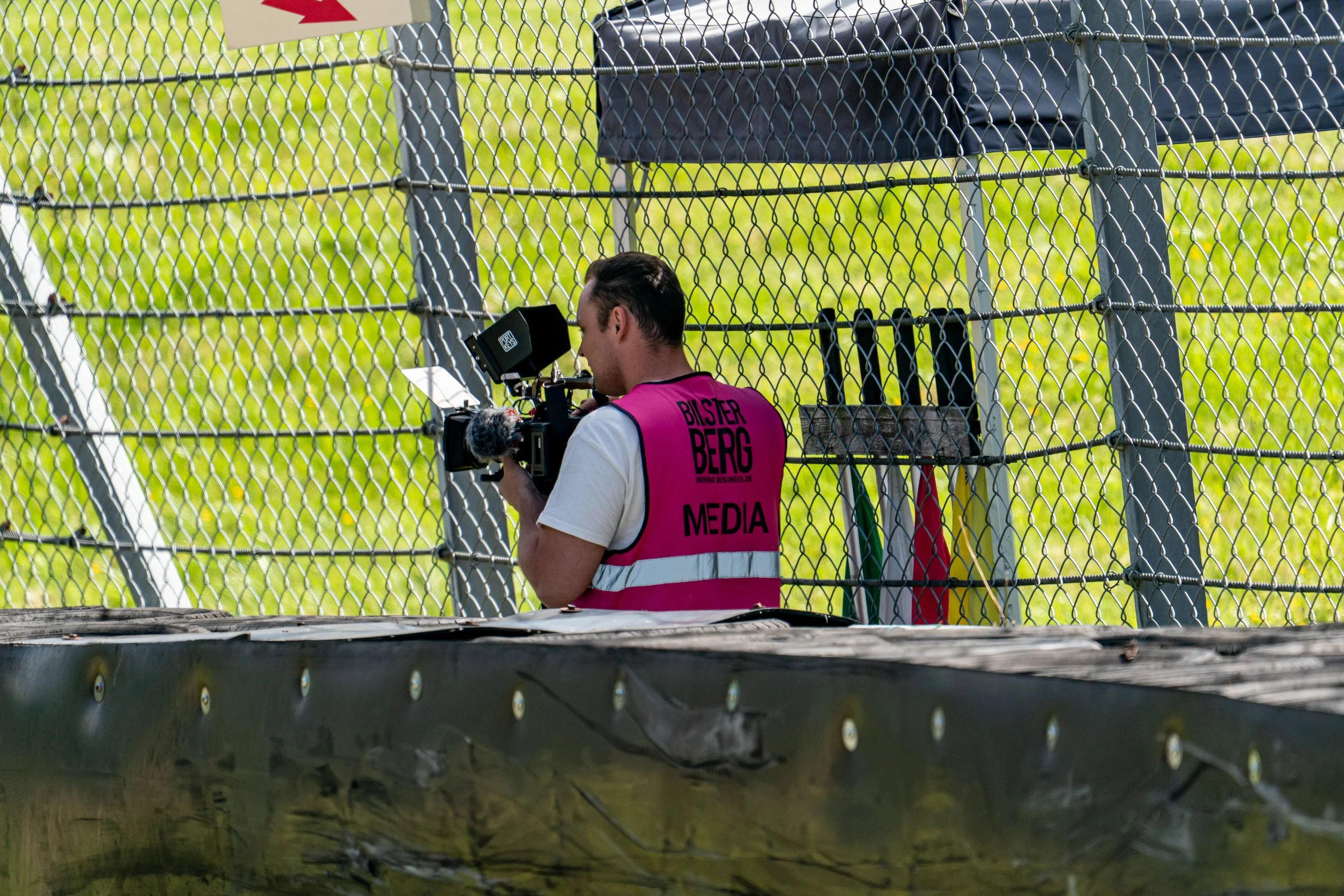 Ein Mann mit einem Videokamerasystem, der vor einem Zaun steht und auf ein Event setzt, getragen von einem pinkfarbenen Sicherheitsweste mit Aufschrift Medien.
