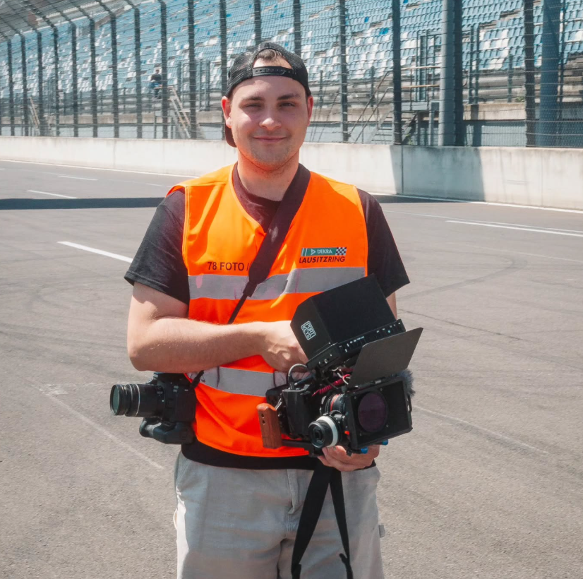 Junger Mann mit Rucksack und professioneller Videokamera auf einer Rennstrecke, trägt eine orange Sicherheitsweste, im Hintergrund leere Tribünen.