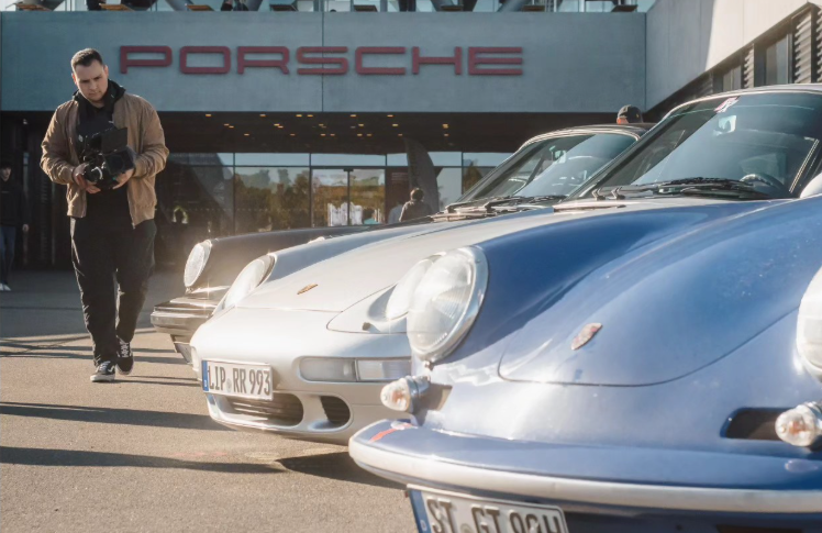 Mehrere klassische Porsche-Sportwagen vor einem Porsche-Händler, mit einem Mann, der eine Kamera hält, im Vordergrund.