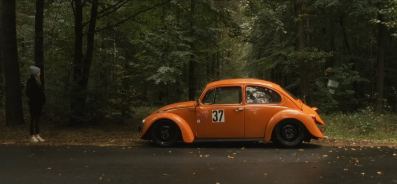 Ein orangefarbener Volkswagen Käfer mit der Nummer 37 parkt am Straßenrand in einem Wald. Links steht eine Person in dunkler Kleidung und einem grauen Hut, die in Richtung des Autos blickt.