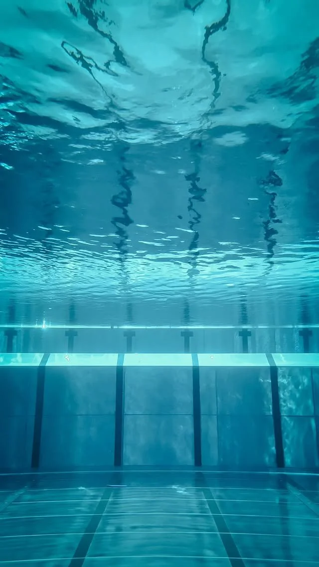 Unterwasseraufnahme eines leeren Schwimmbeckens mit dunklen Linien auf dem Boden und Wasseroberfläche mit Reflexionen.