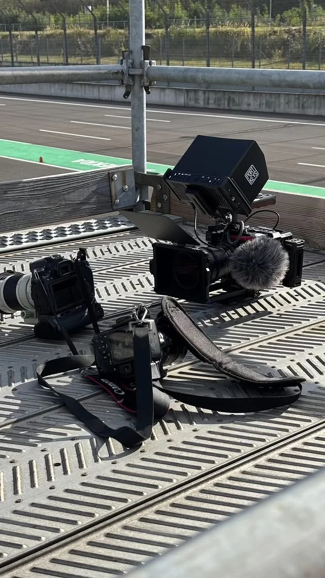 Fotografie von mehreren Kameras und Aufnahmegeräten auf einer Tribüne, wahrscheinlich am Rennstrecke, mit Blick auf eine leere Rennstrecke im Hintergrund.
