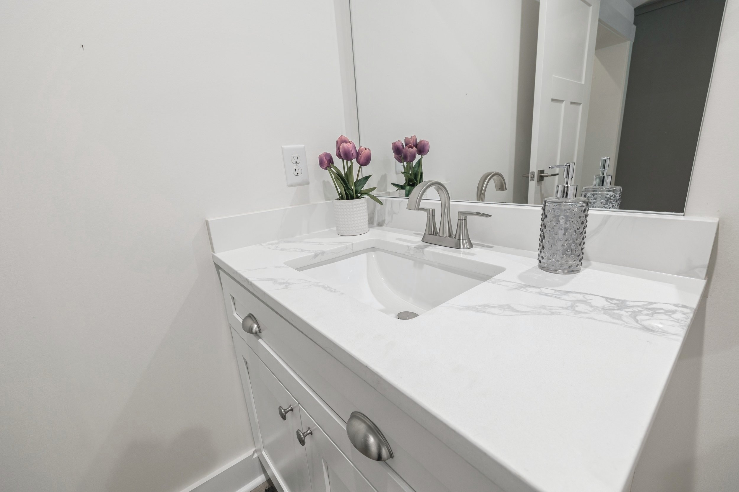 Modern bathroom vanity with white countertop, a sink, silver faucet, mirrored backsplash, potted purple flowers, and a textured soap dispenser.