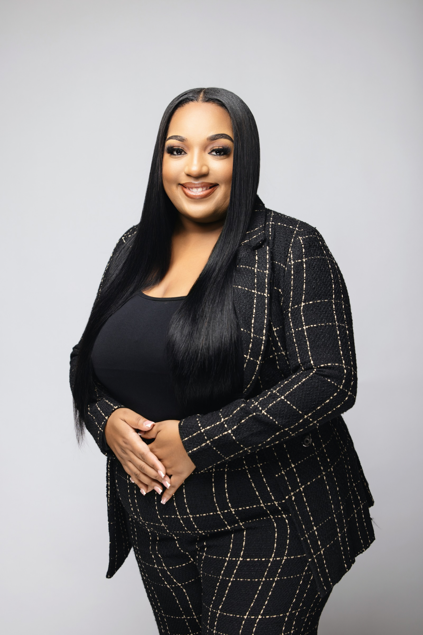 A smiling woman with long, straight black hair wearing a black top and a black blazer and pants with a checkered pattern, posing against a light gray background.