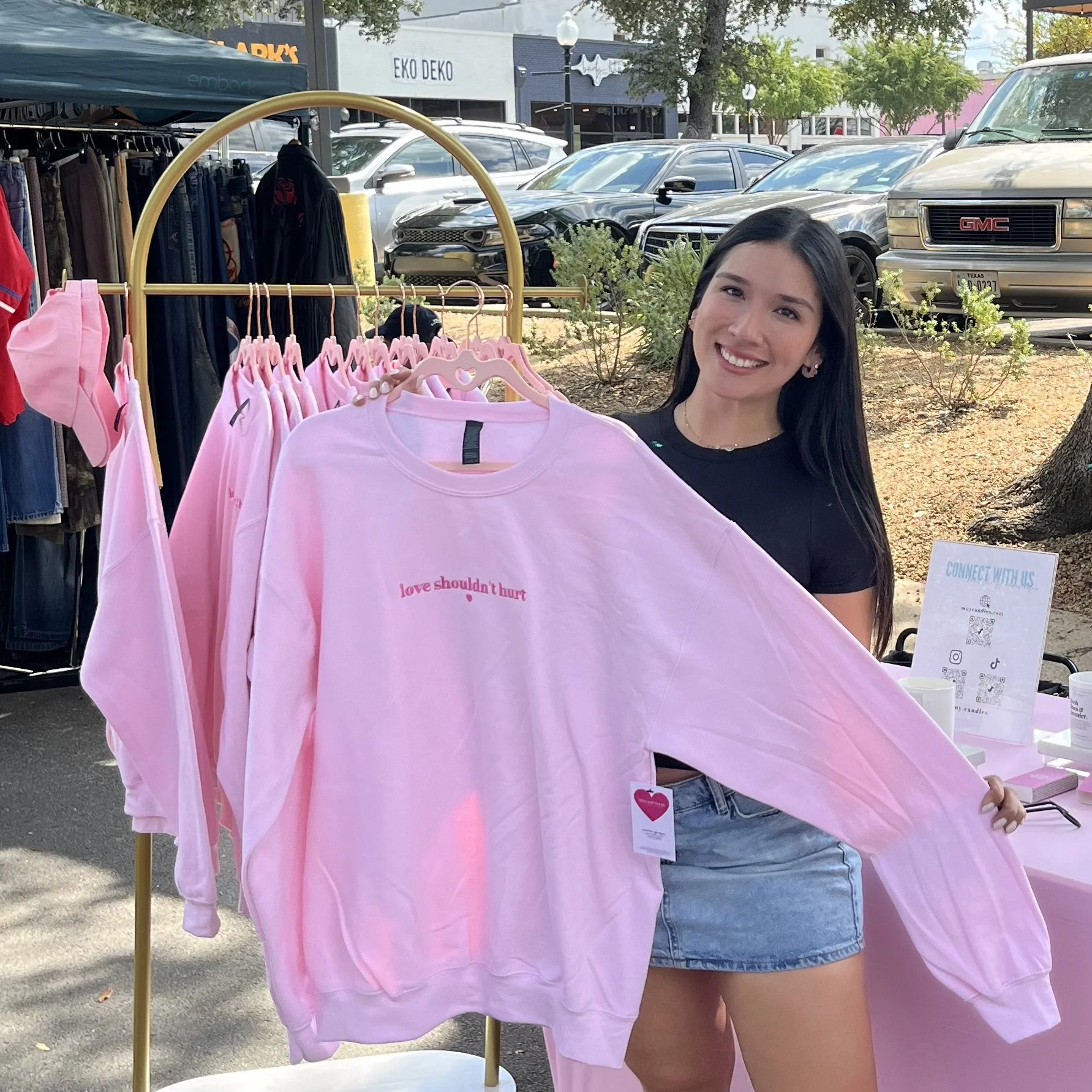 A smiling woman standing outdoors next to a pink sweatshirt on a clothing rack. The sweatshirt has the words "love shouldn’t hurt" written on it. There are cars and trees in the background.