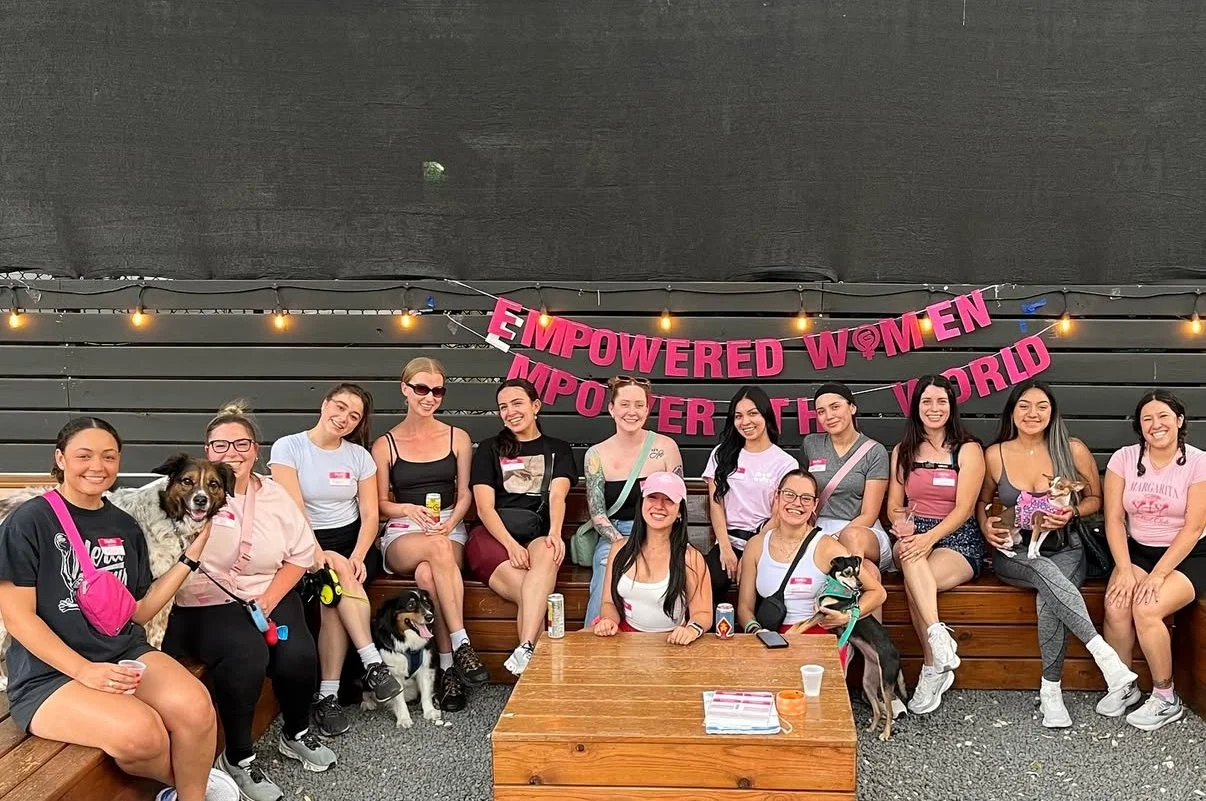 Group of women sitting and standing together under a pink banner that reads 'Empowered Women Empower the World,' with string lights overhead. Some women are holding dogs and drinks, smiling for the photo.