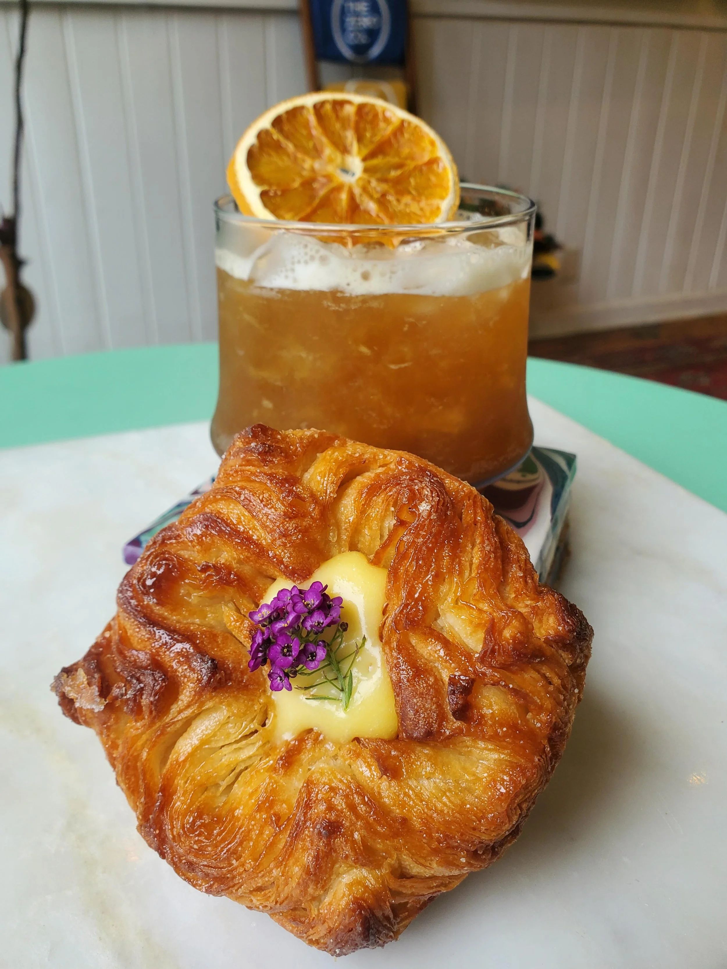 Flaky laminated pastry with lemon curd and purple flowers alongside a craft cocktail with dried citrus at Colette Bread & Bakeshop Atlanta