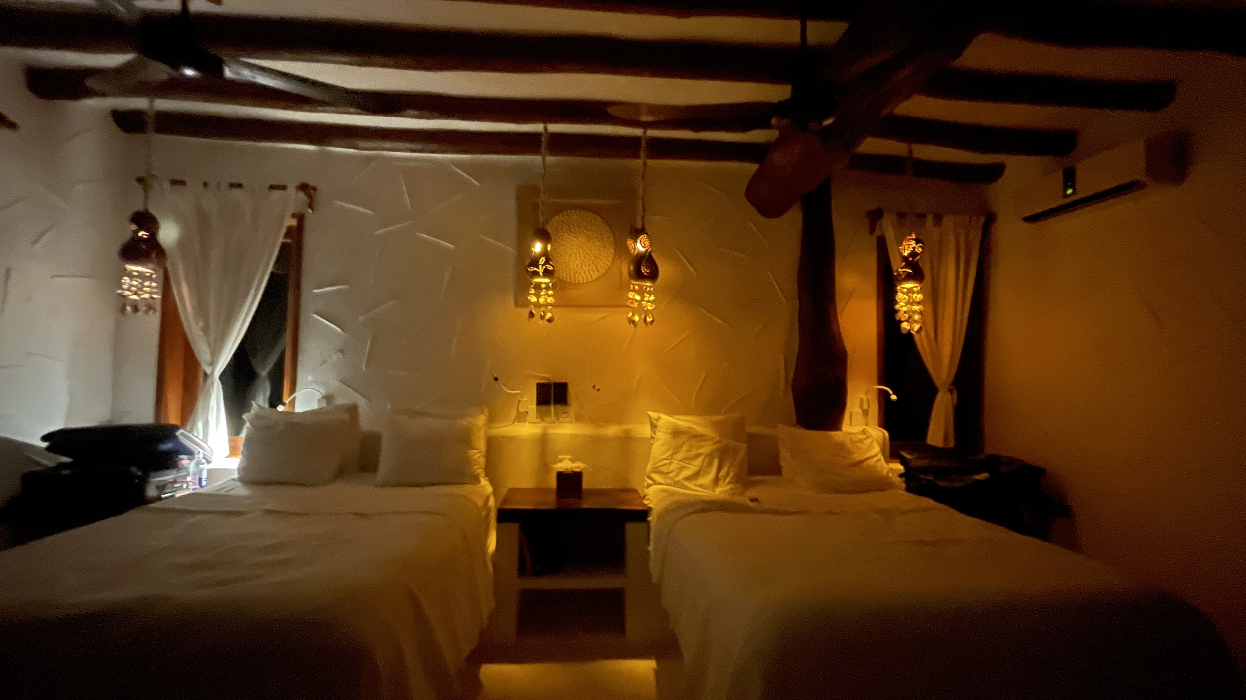 A cozy hotel room with two beds, each with white bedding and pillows, warm yellow lighting, wooden beams on the ceiling, two small windows with curtains, and decorative hanging lamps.